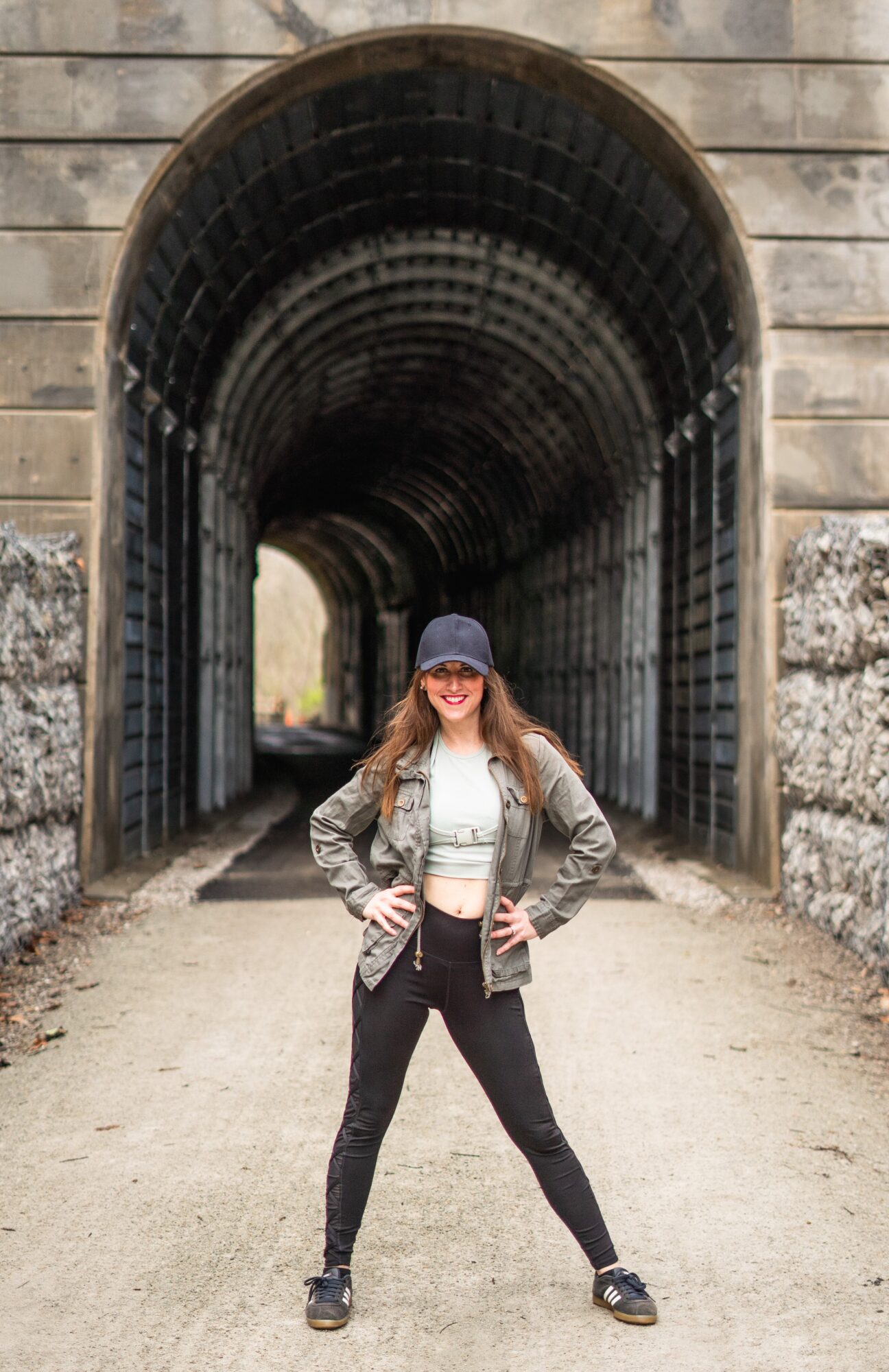 Woman standing with hands on hips in front of a tunnel entrance, smiling, wearing a cap, jacket, and sunglasses.
