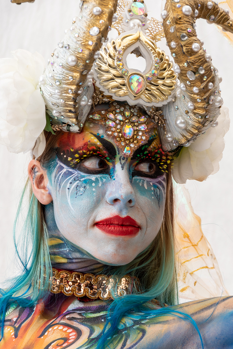 Person with elaborate headdress, colorful makeup, and face paint, wearing ornate jewelry and costume, looking directly at camera.