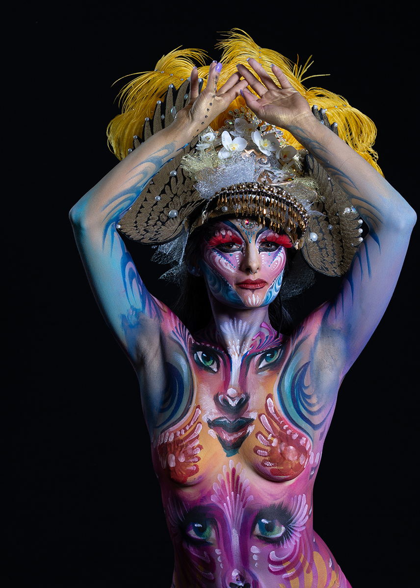 Person with elaborate headpiece and body paint featuring multiple faces and vibrant colors, posing with hands on head.