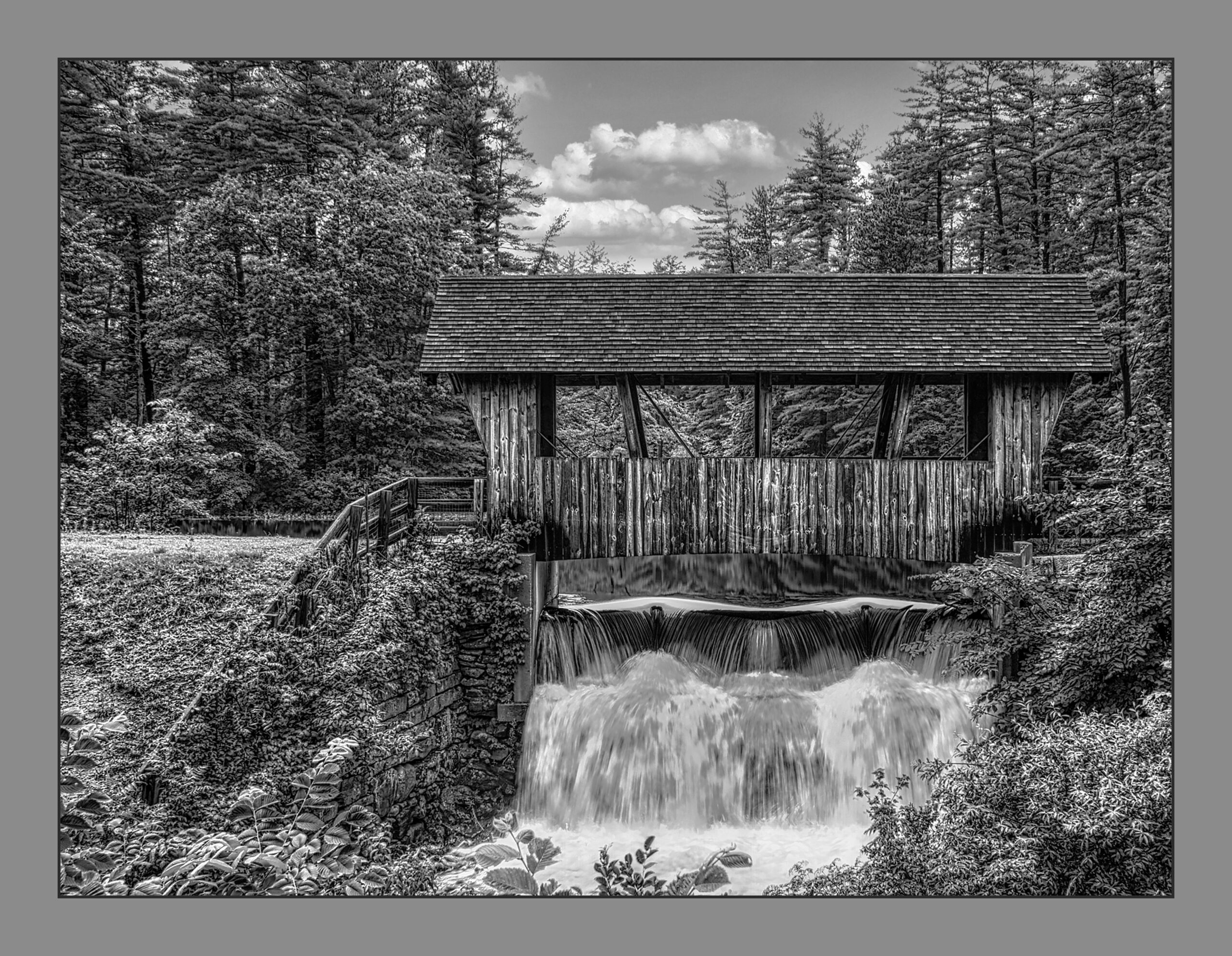 A black and white photo of a covered wooden bridge over a small waterfall in a forested area.