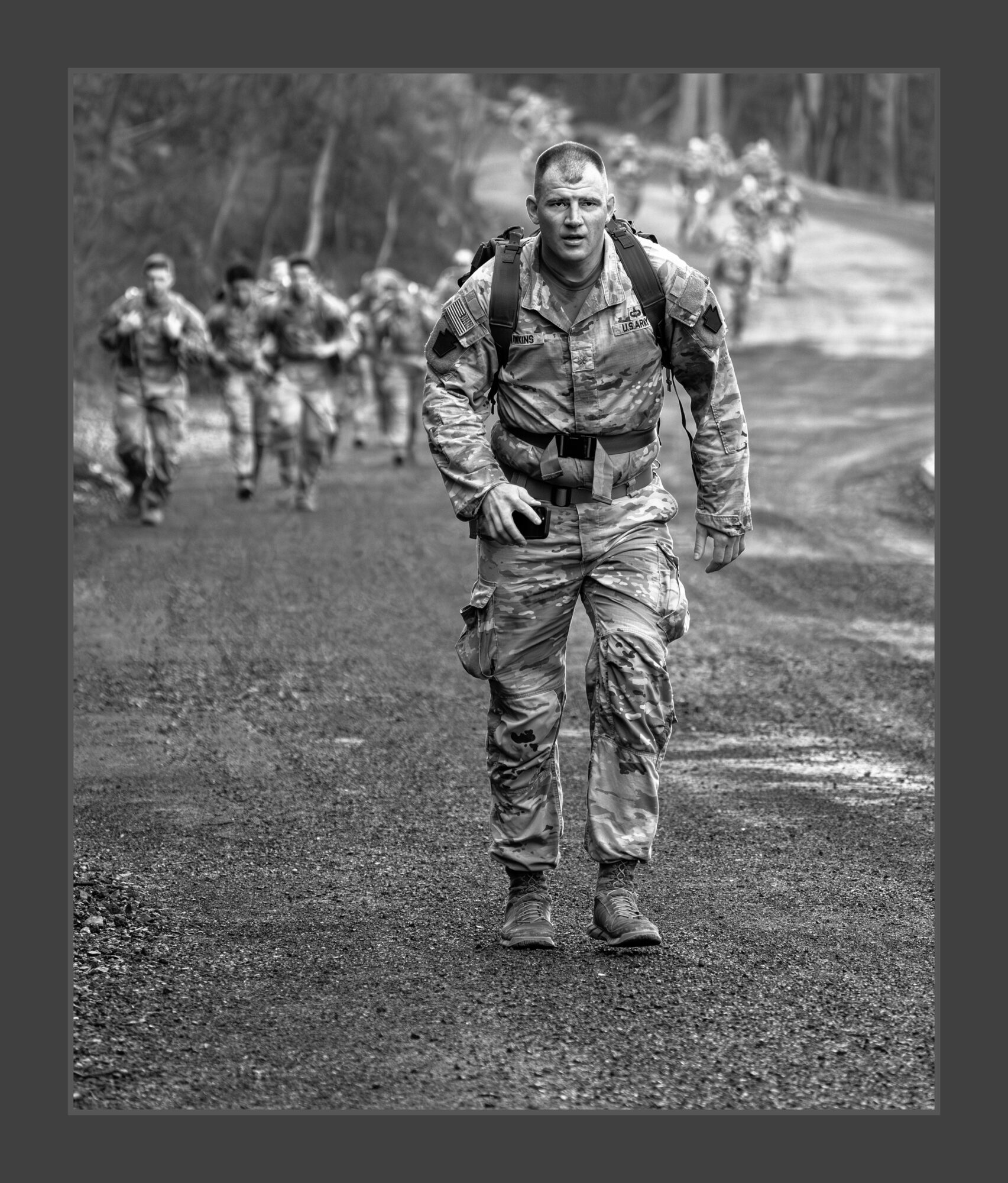 Soldier walking on a dirt road with a group of soldiers in the background, black and white image.