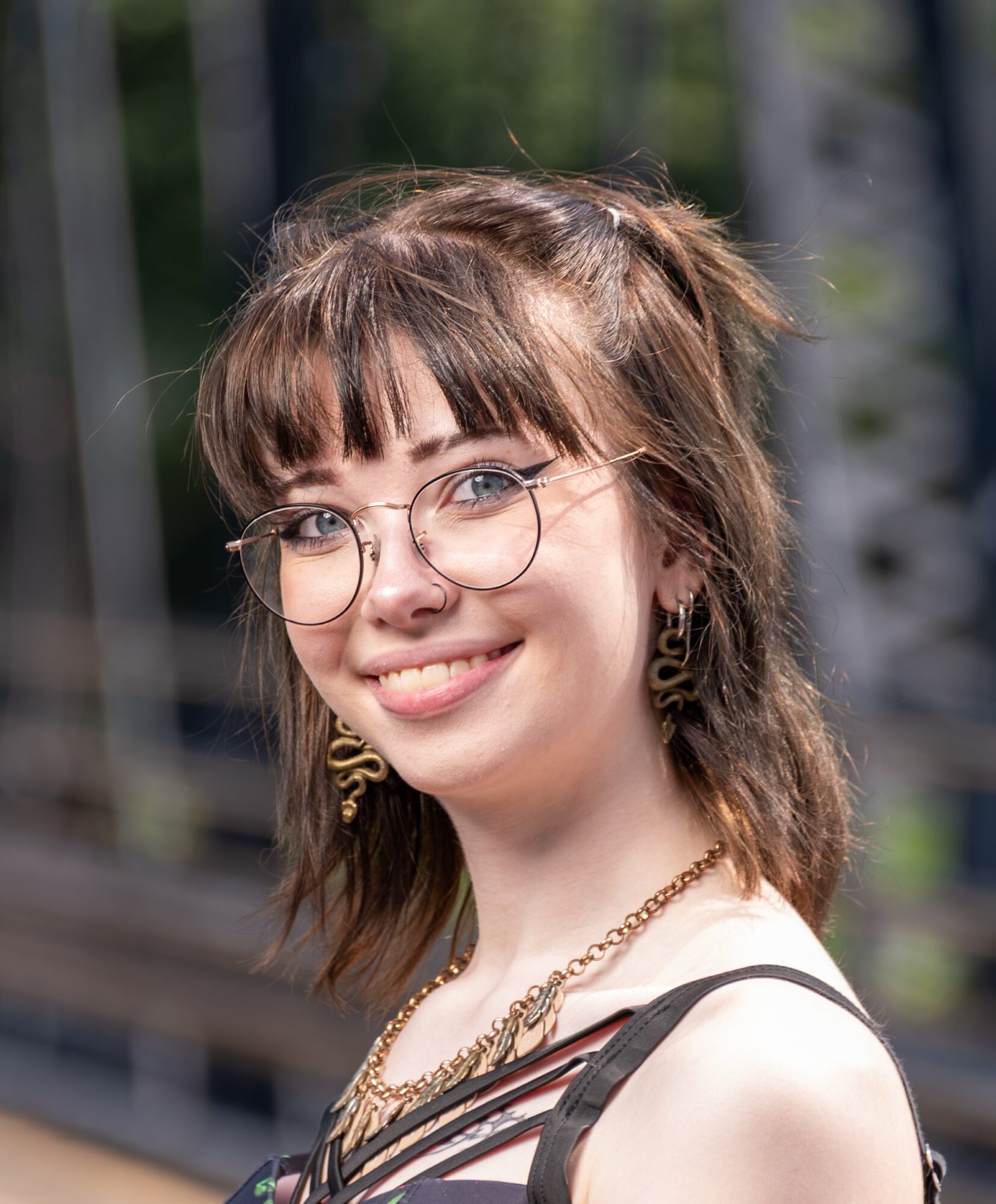 Young woman with glasses, earrings, and layered necklaces smiling outdoors, with blurred background of trees and structures.