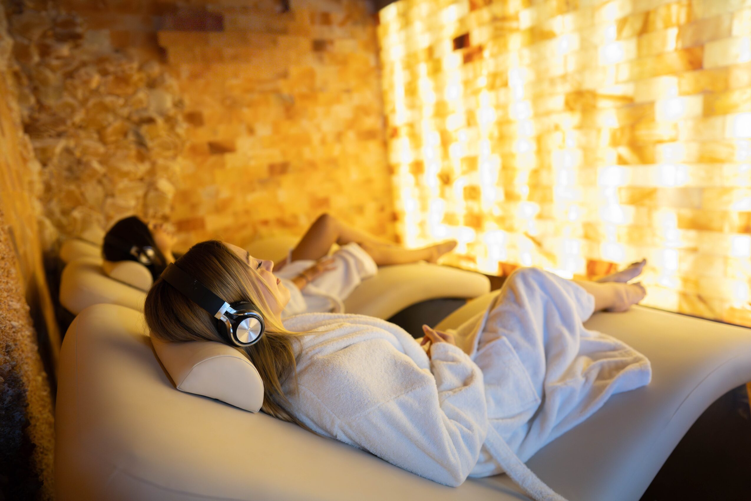 Person lying on a lounge chair with headphones, relaxing in a room with warm lighting and brick wall background.