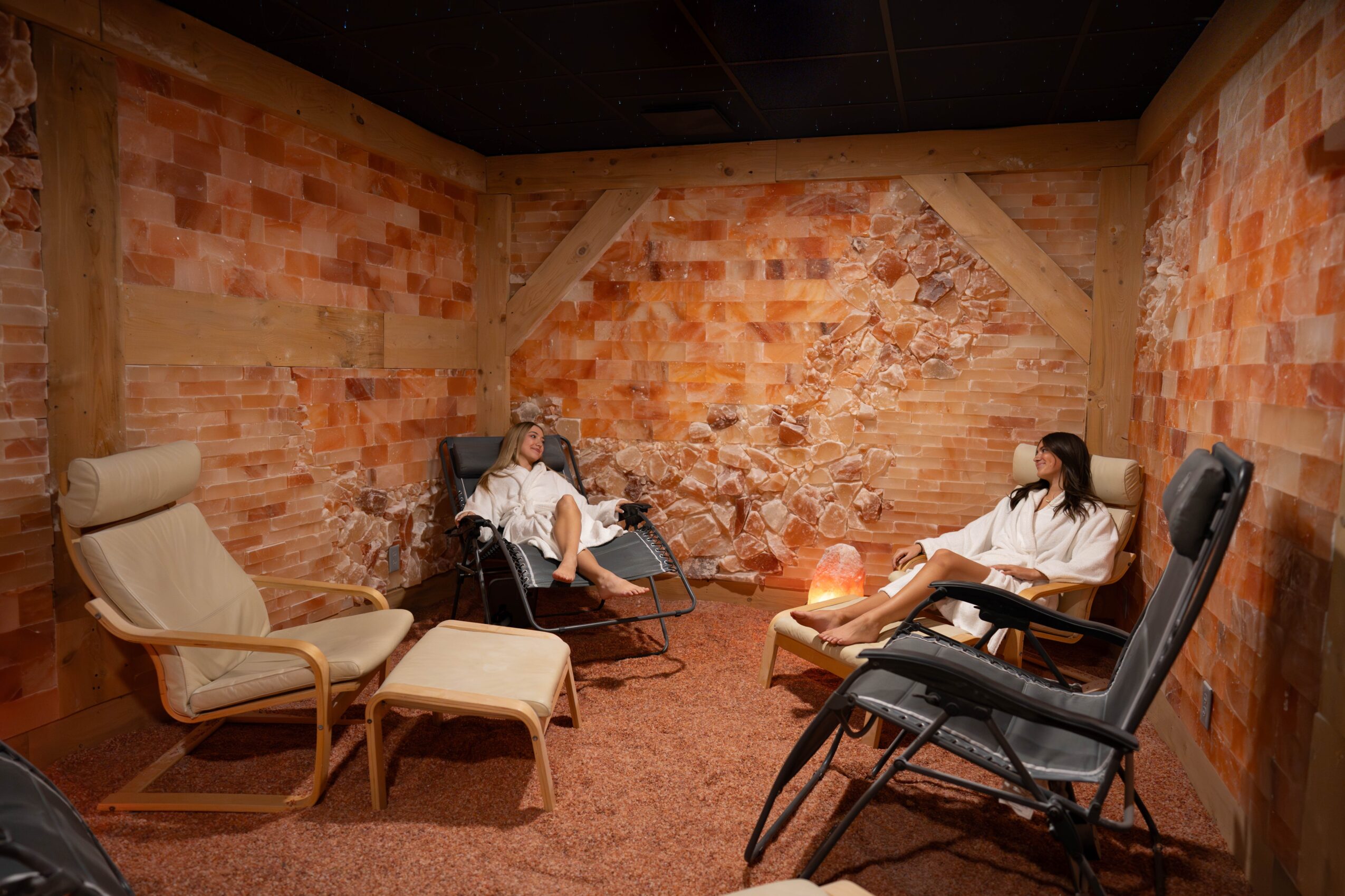 Two people sit in a room with brick walls, one on a beige chair and the other on a black chair, with a small table between them.