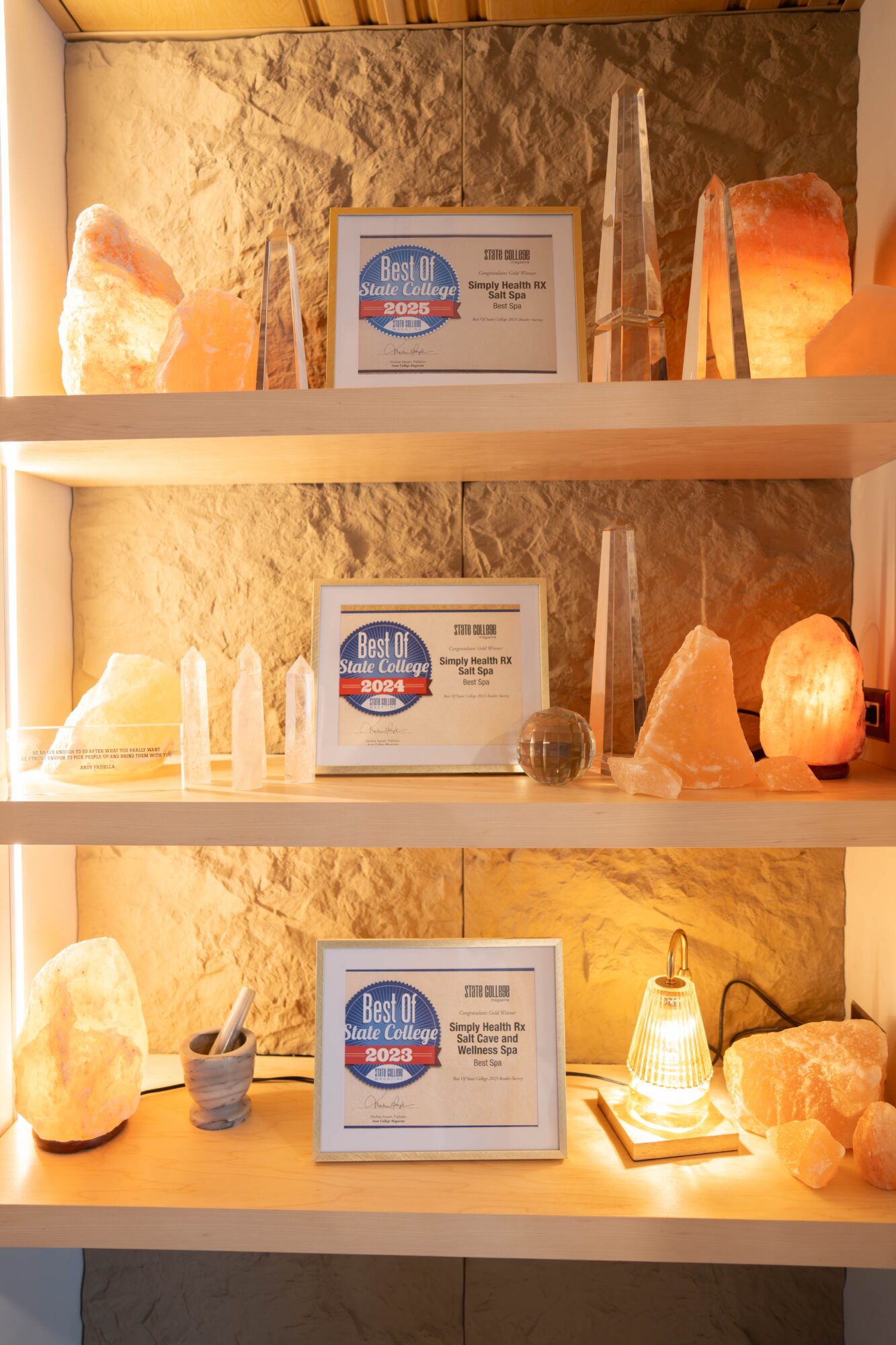 Three shelves with salt lamps, salt crystals, and framed signs, illuminated by warm lighting, against a textured wall.