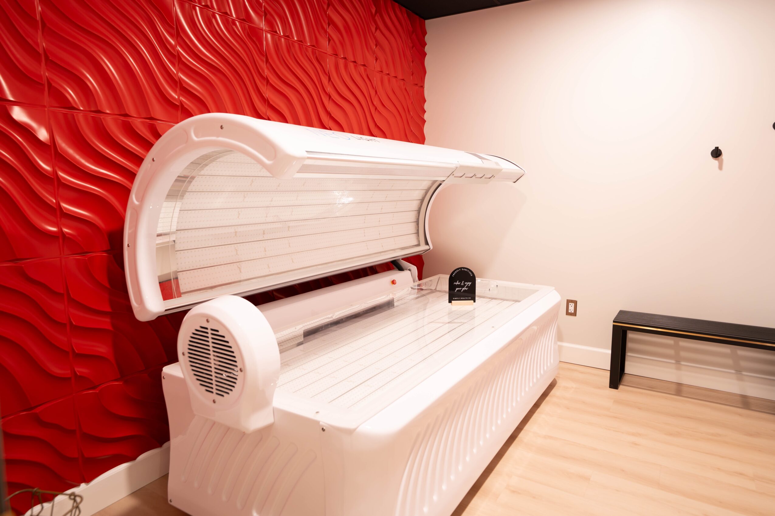 Indoor tanning bed with white exterior and open lid, against a red textured wall and white wall, with a black bench nearby.