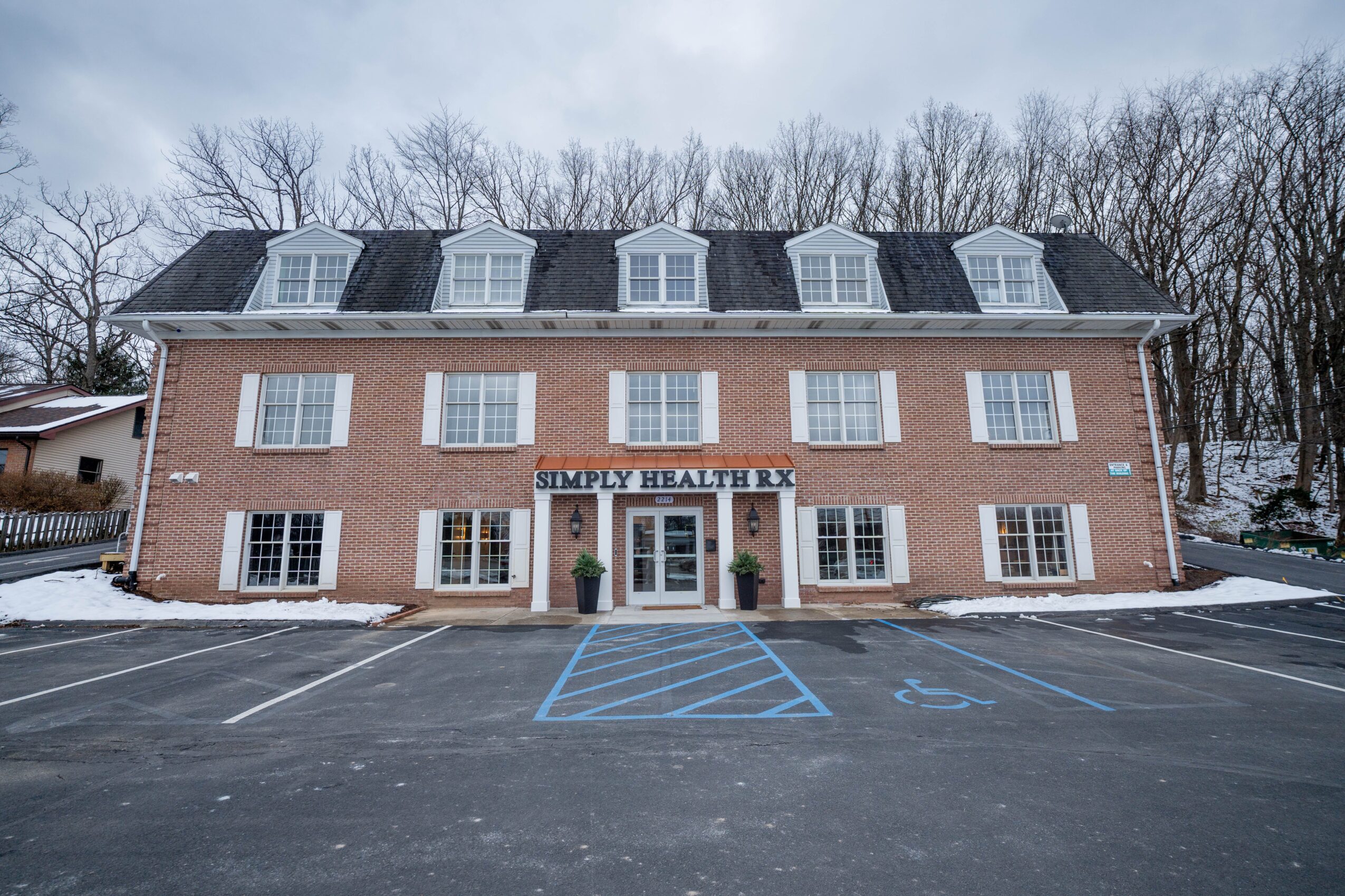Three-story brick building with a sign reading 'Simply Health' above the entrance, surrounded by a parking lot and leafless trees.