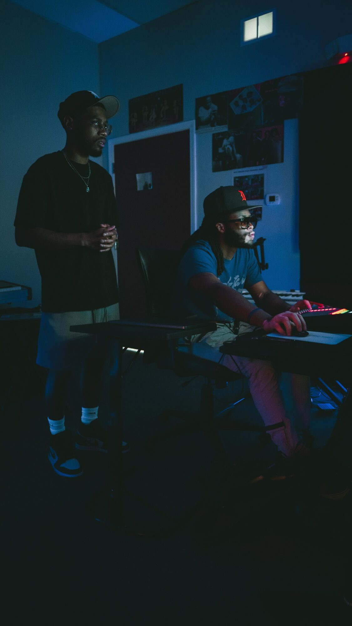 Two people in a dark room with electronic equipment, one standing and one sitting at a table, illuminated by screen light.