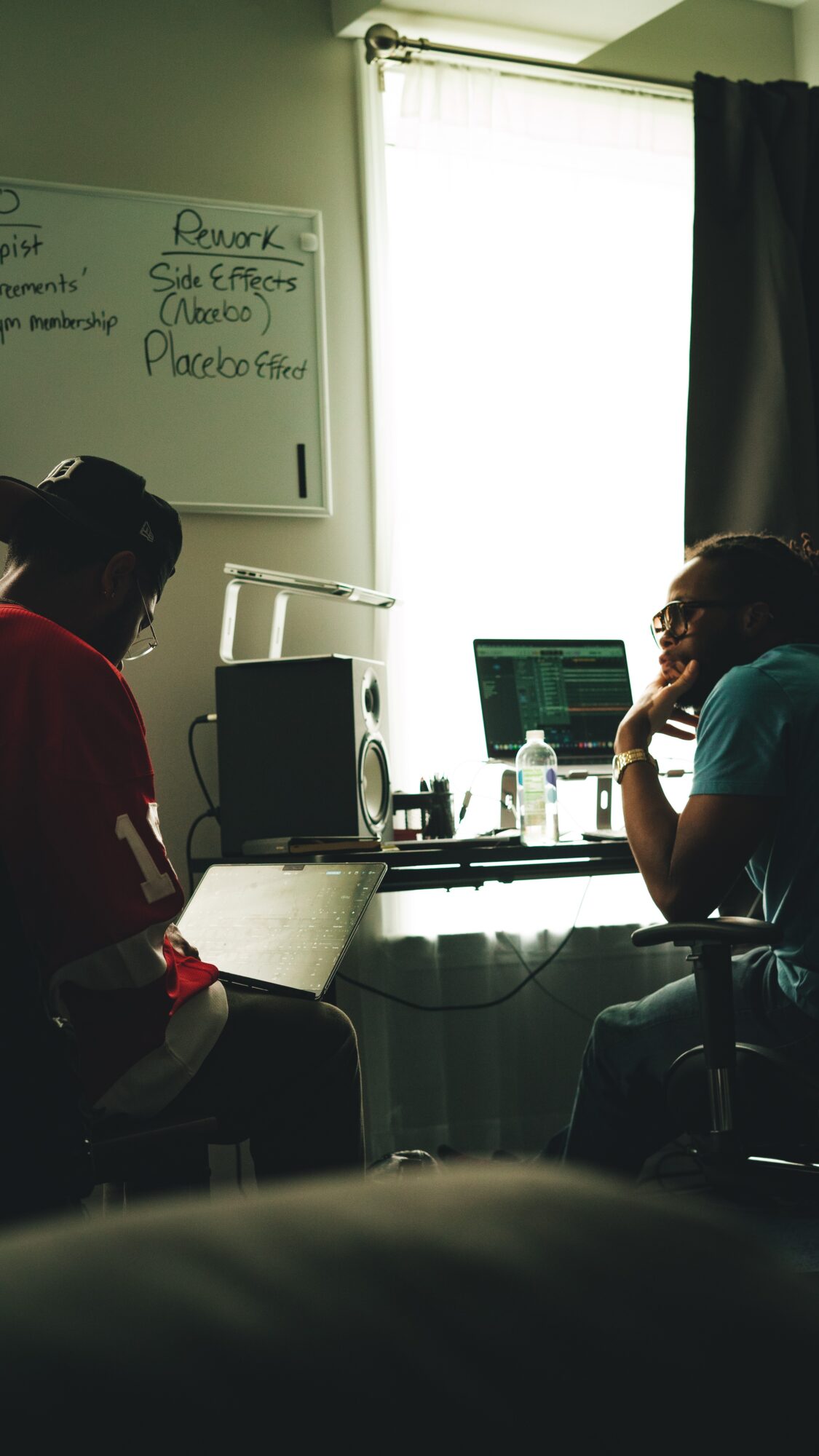 Two people sitting at a desk with computers, a whiteboard, and a window with curtains behind them.