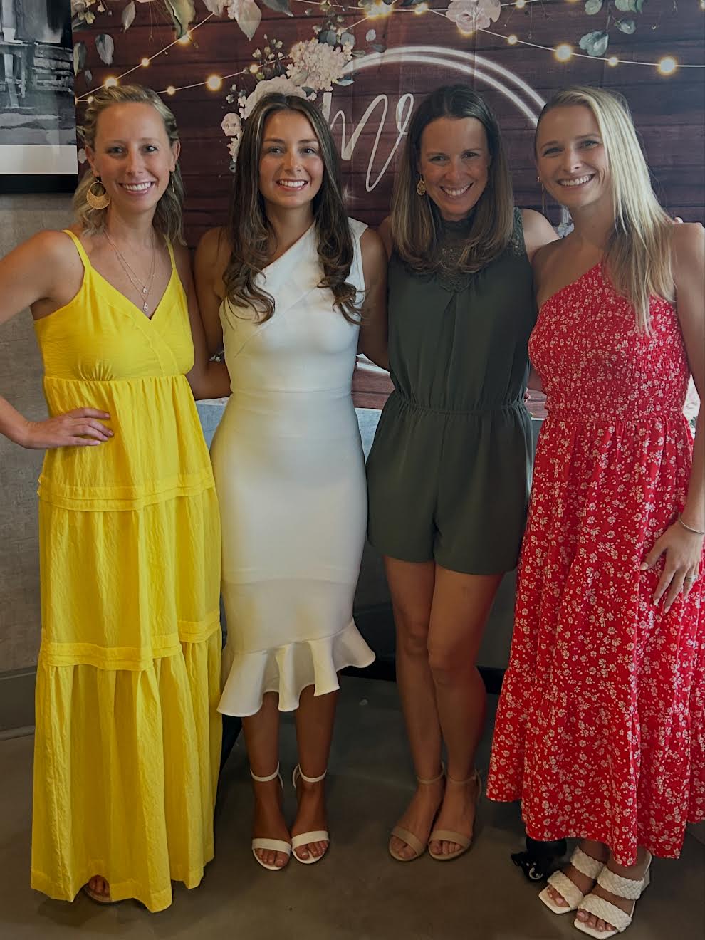 Four women standing together, smiling, in front of a decorated backdrop with string lights and a heart-shaped sign.