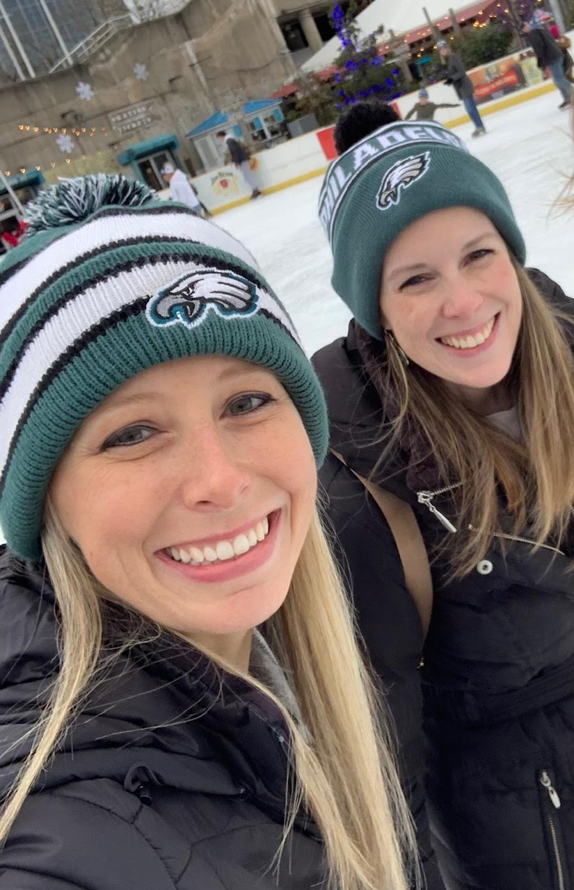 Two smiling women wearing Philadelphia Eagles beanies outdoors on ice rink, winter clothing, with ice and people in background.