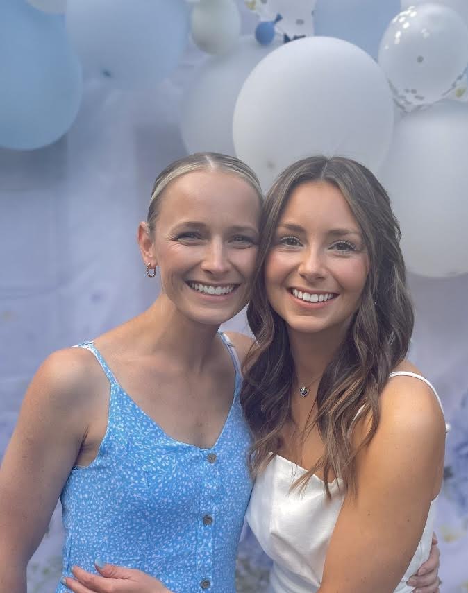 Two smiling women with long hair, one in a blue dress and the other in a white dress, standing close together.