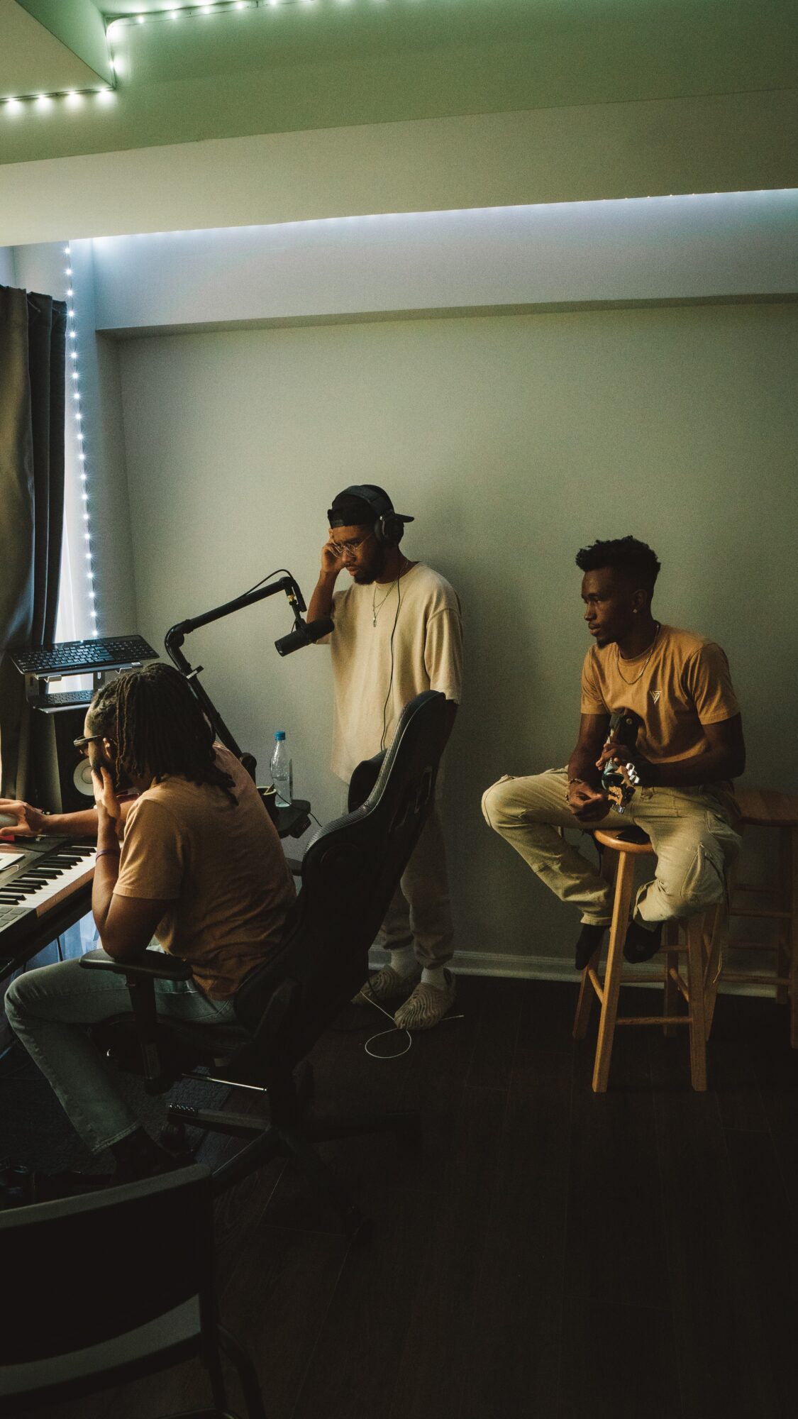 Three people in a room with musical instruments and a microphone, one person singing, others playing guitar and keyboard.
