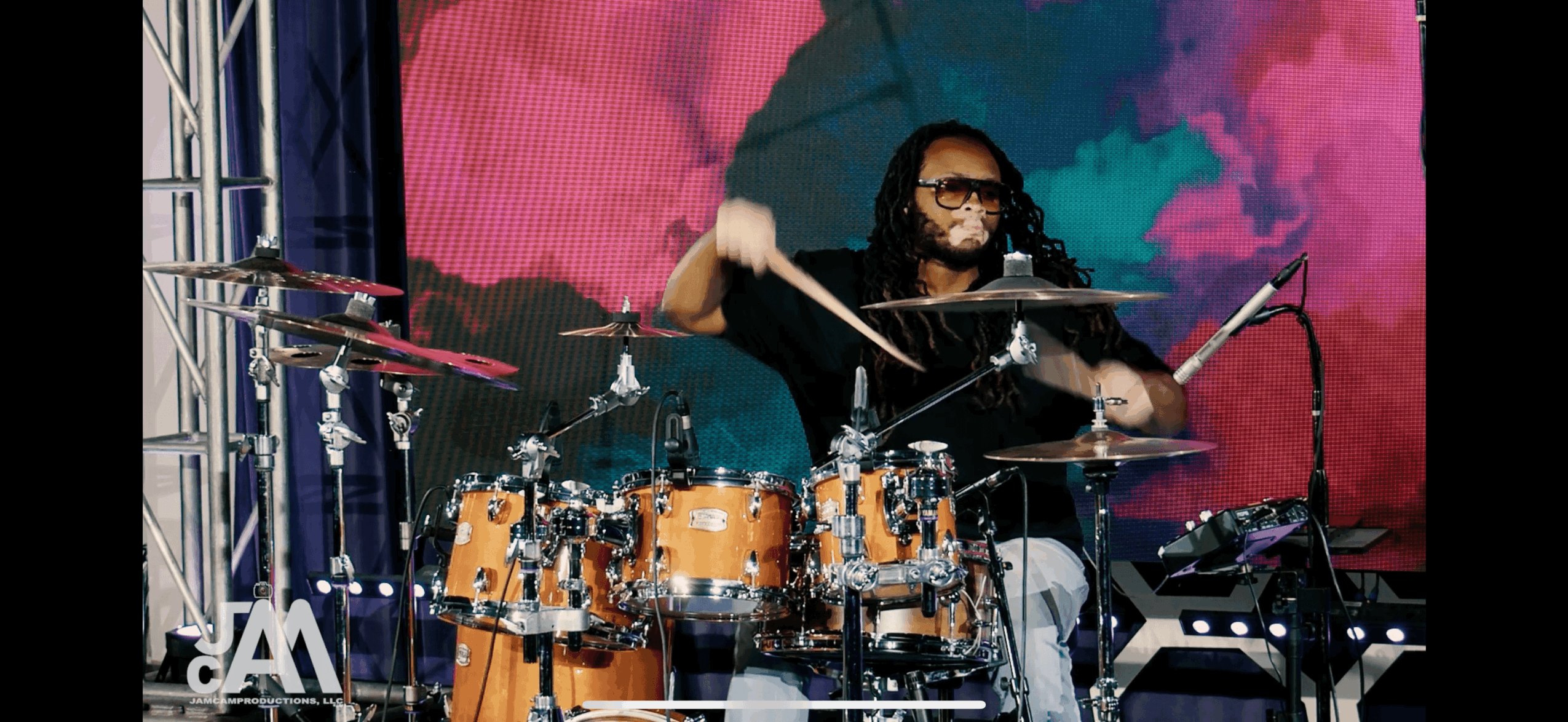 Person playing drums on stage with colorful background, wearing glasses and dark clothing.