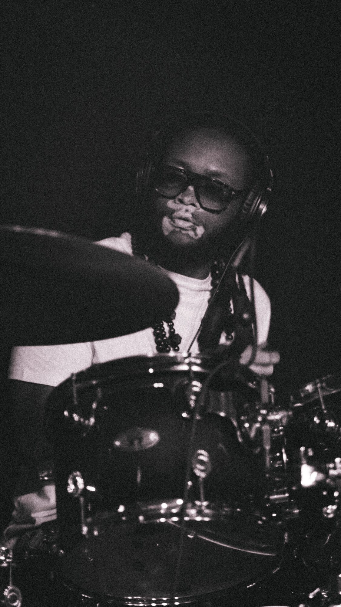 Person with glasses playing drums in a dark setting, wearing headphones, with drum kit visible.