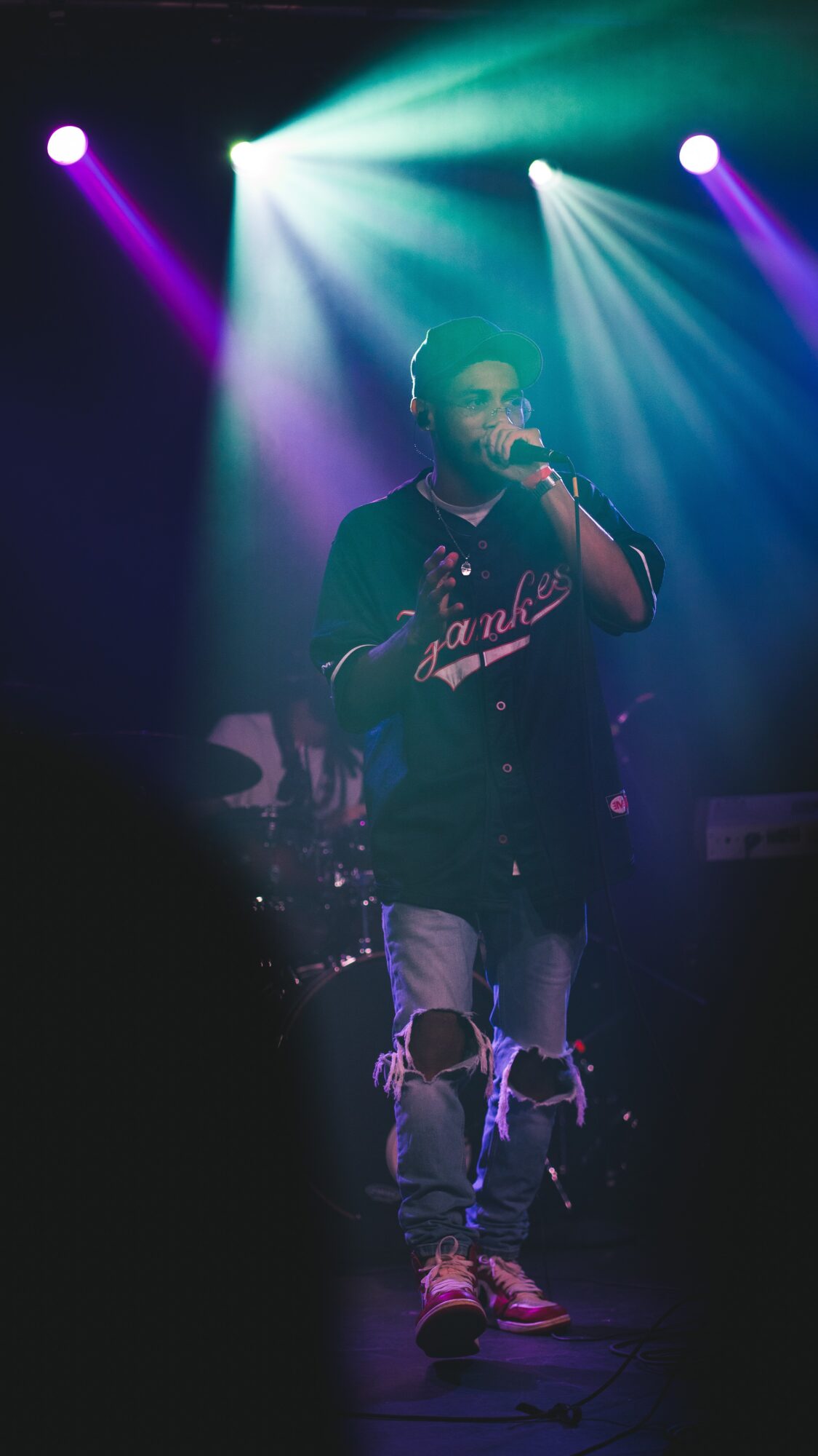 Performer singing into a microphone on stage with colorful lights shining behind, wearing a black jacket and ripped jeans.