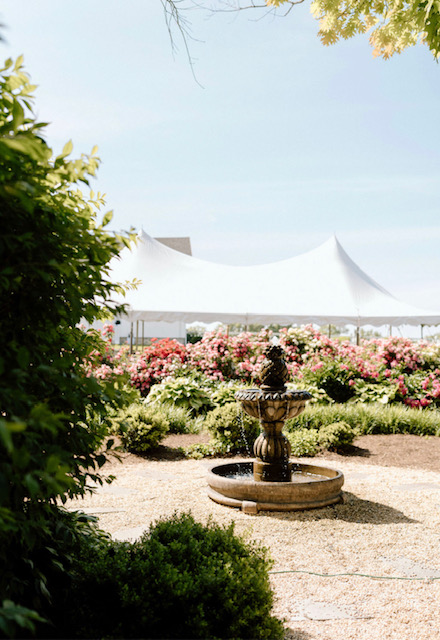 Garden with a fountain, flower beds, and a large white tent in the background, framed by trees and bushes.