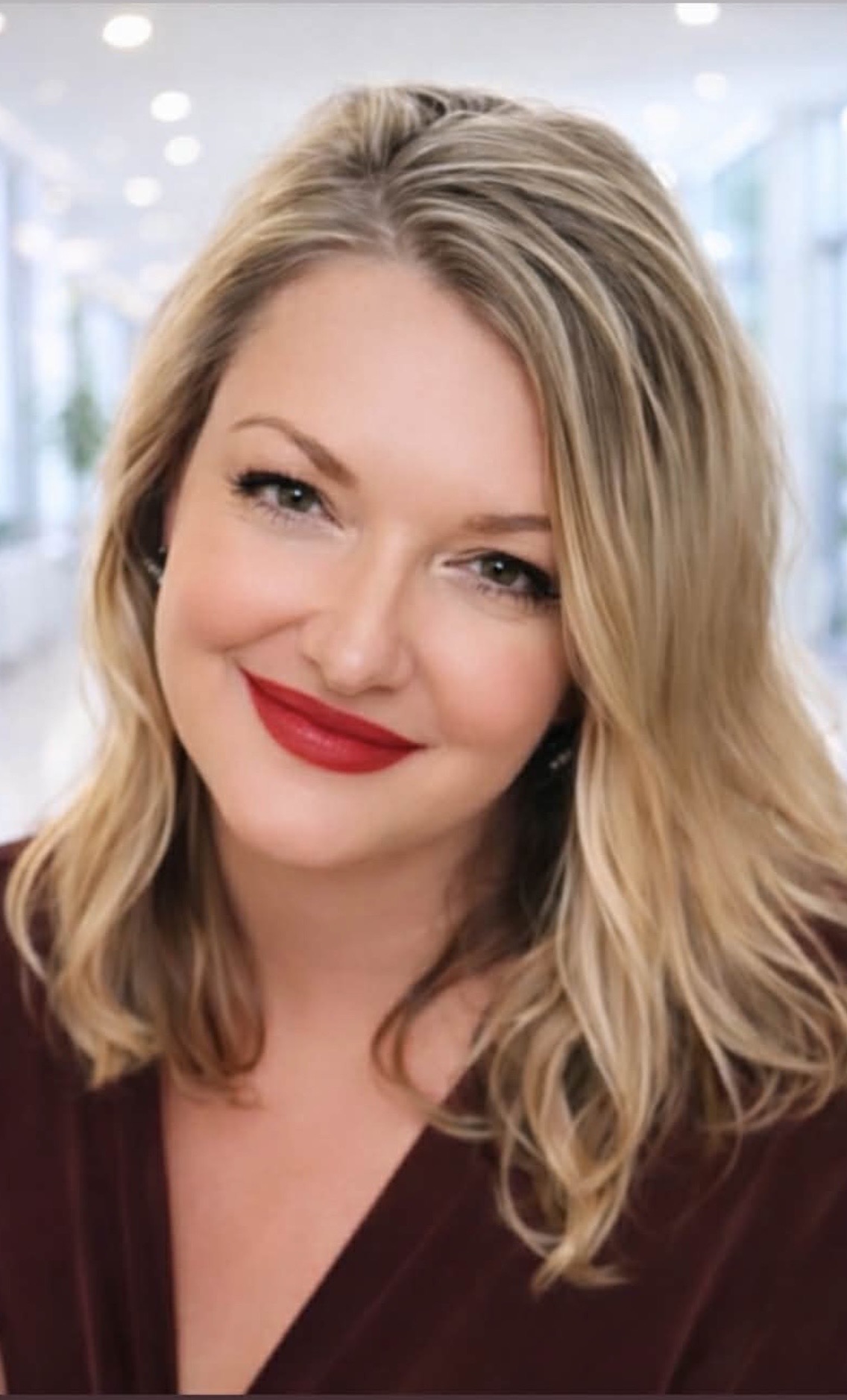 Woman with blonde hair and red lipstick smiling, indoors with bright lighting and blurred background.