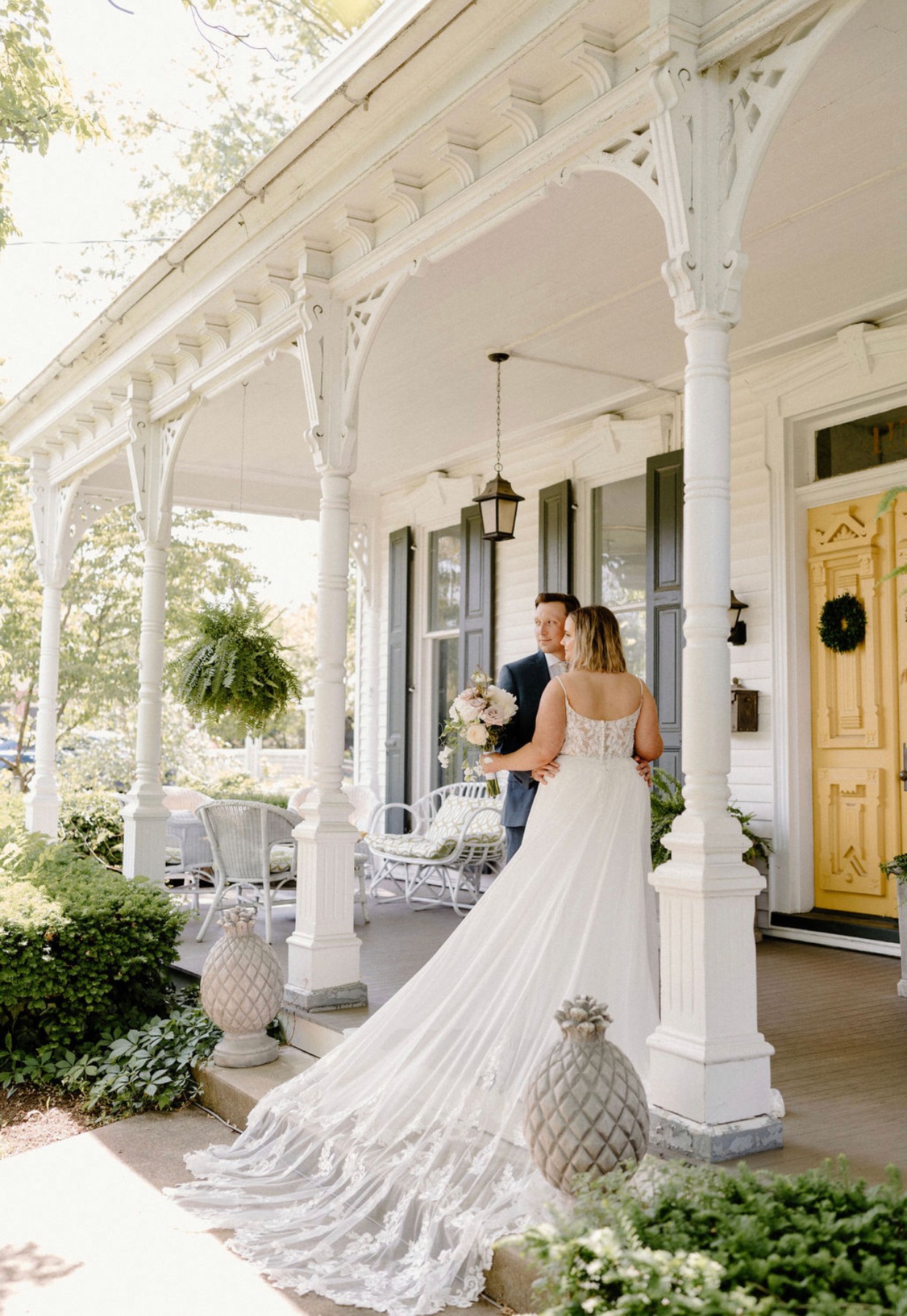 Two women in wedding dresses embrace on a porch with white columns and hanging plants, sunlight filtering through trees.