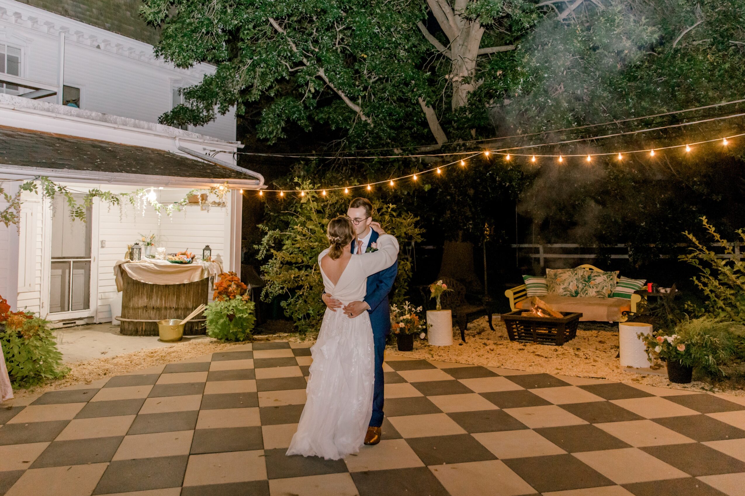 Couple dancing outdoors at night under string lights, with a decorated house and garden in the background.