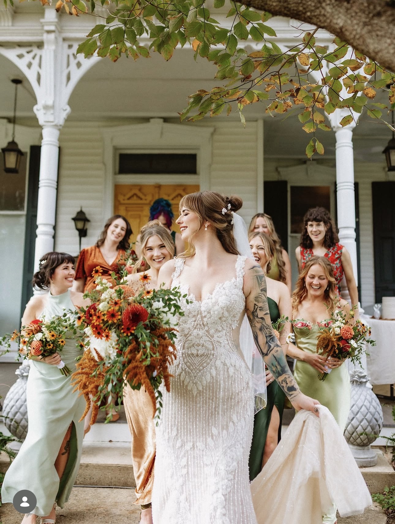 Bride in white dress holding a large bouquet, surrounded by smiling women holding smaller bouquets, on porch with white columns.
