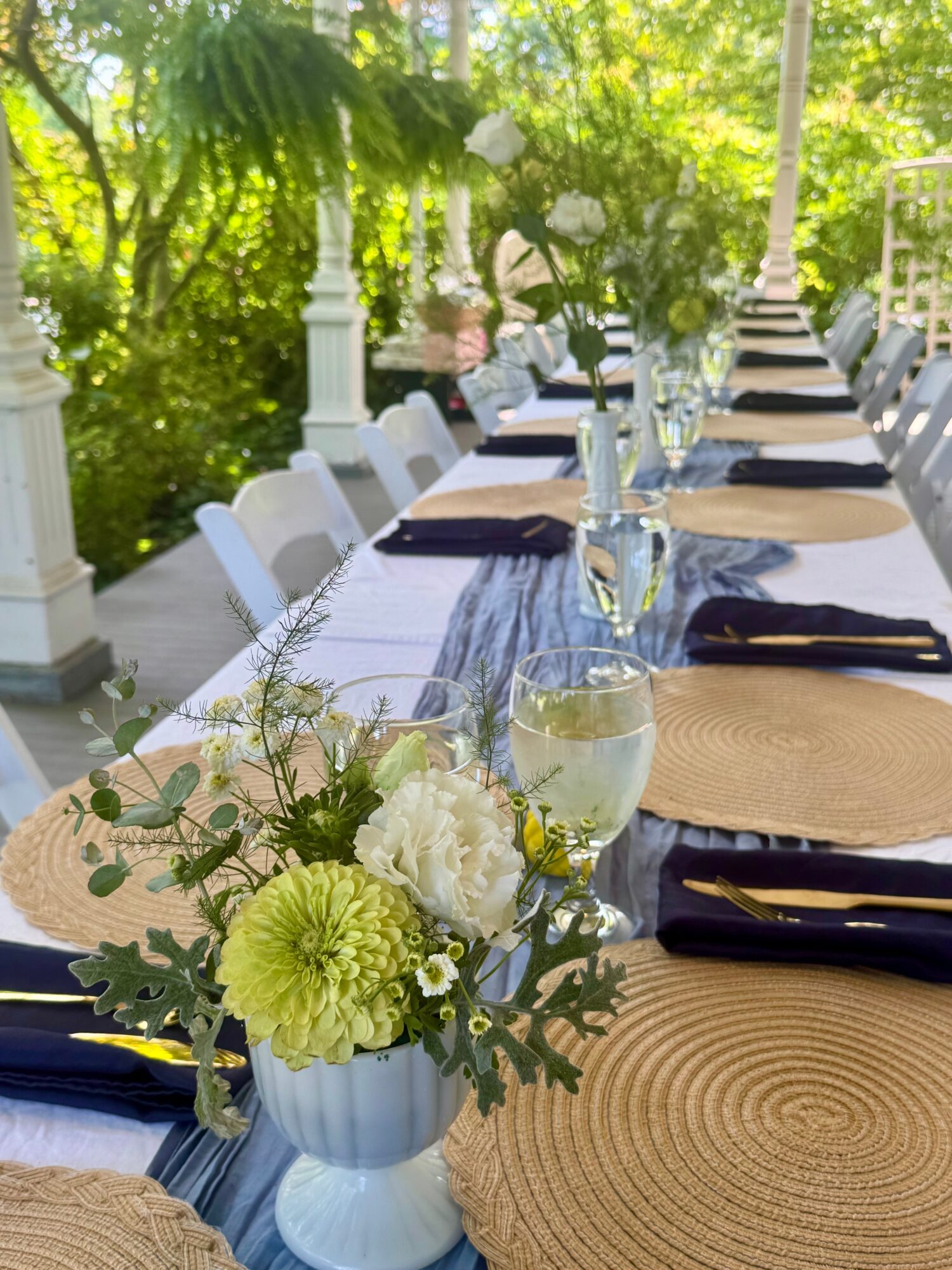 Long outdoor table set with placemats, napkins, glasses, and floral centerpiece, surrounded by greenery and white pillars.