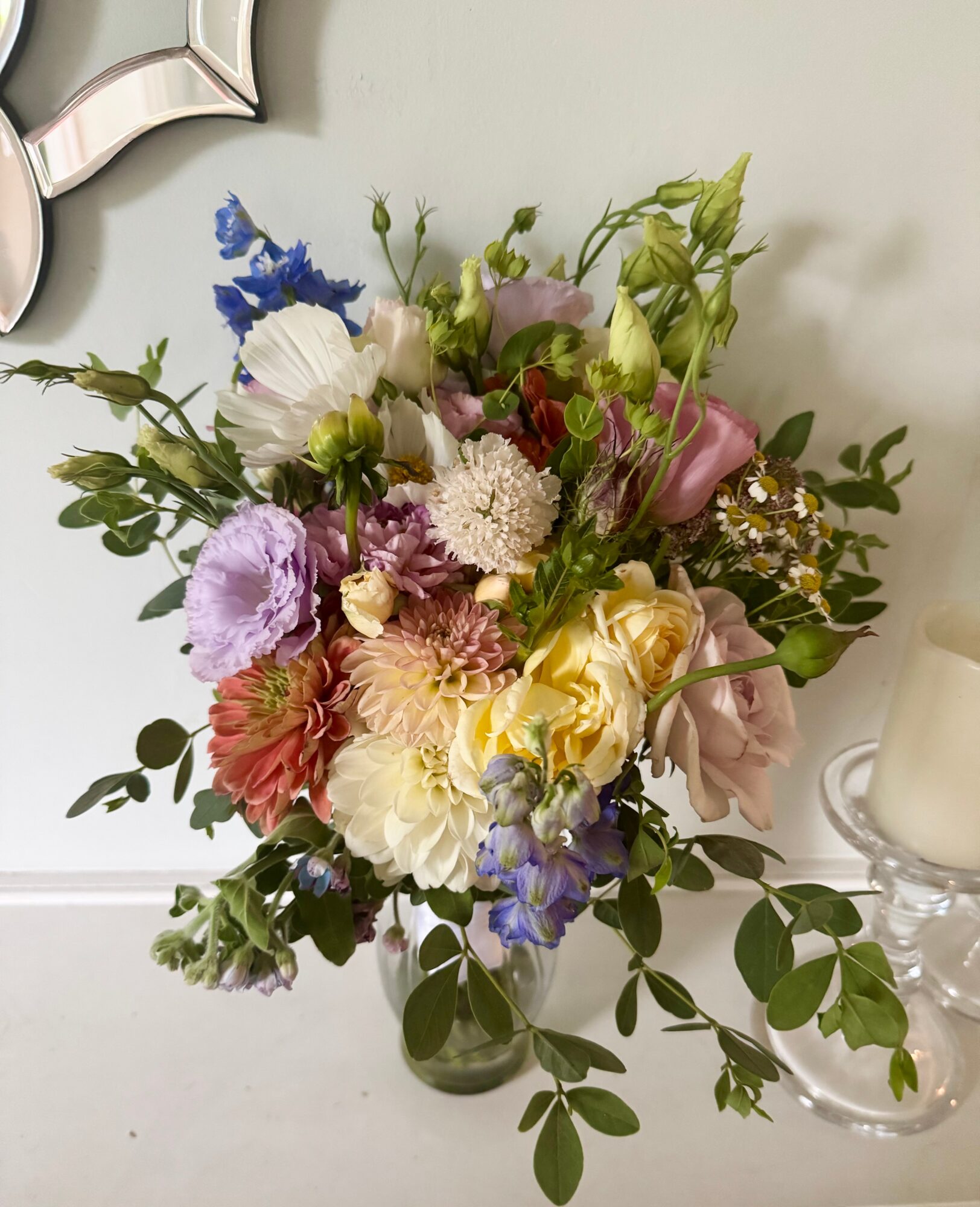 A colorful flower bouquet with various flowers and green leaves in a vase, placed on a white surface.