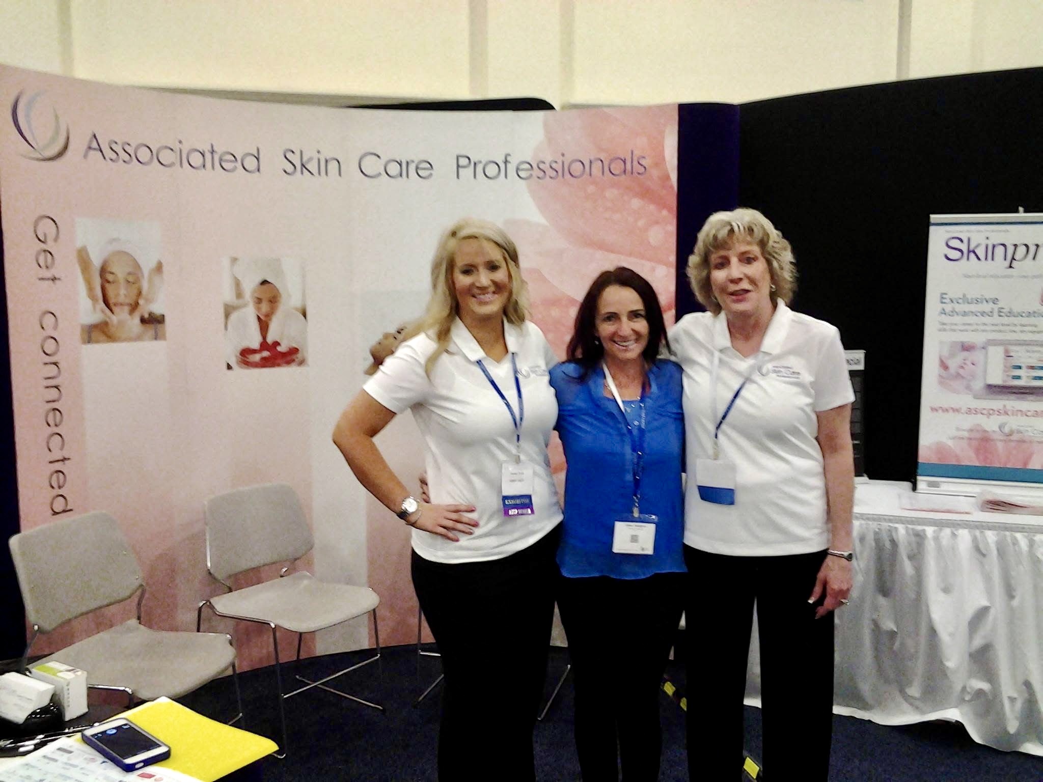 Three women standing together at a skincare professional event, smiling, with a banner and display in the background.