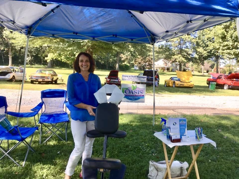 Woman standing under a blue canopy, holding papers, with chairs and a table with items nearby, outdoors on a sunny day.