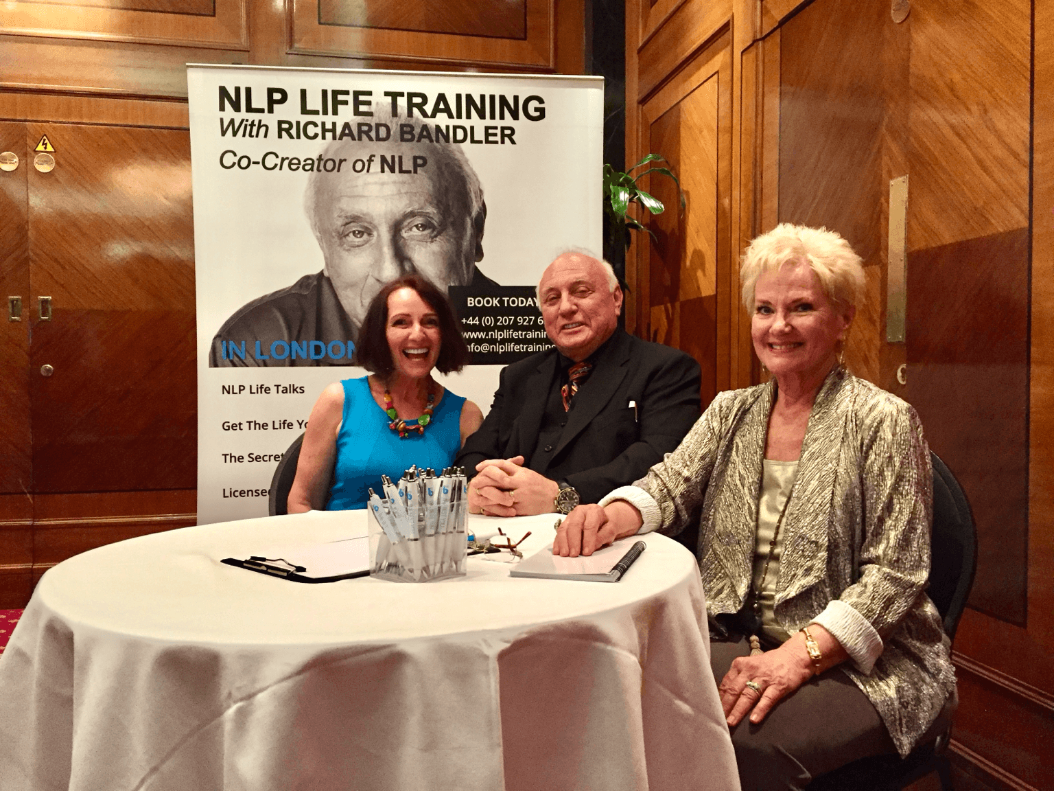 Three people sit at a round table with a banner behind them reading 'NLP Life Training with Richard Bandler.'