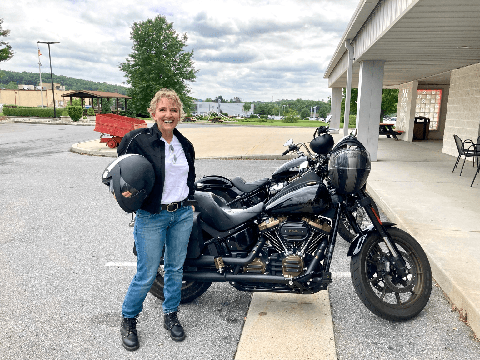 Smiling woman standing next to a black motorcycle outside a building, holding a helmet, with a parking lot and trees in background.