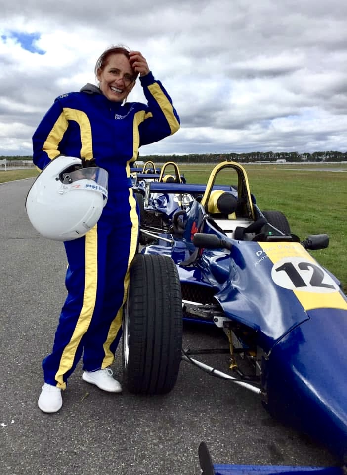 Person in racing suit holding helmet, standing next to a blue race car on track, smiling, with open sky and grass background.