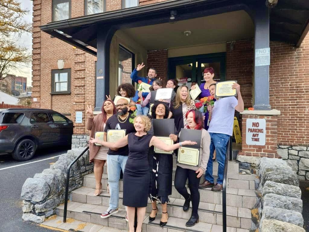Group of people on steps holding certificates, smiling, with a brick building and cars in background.