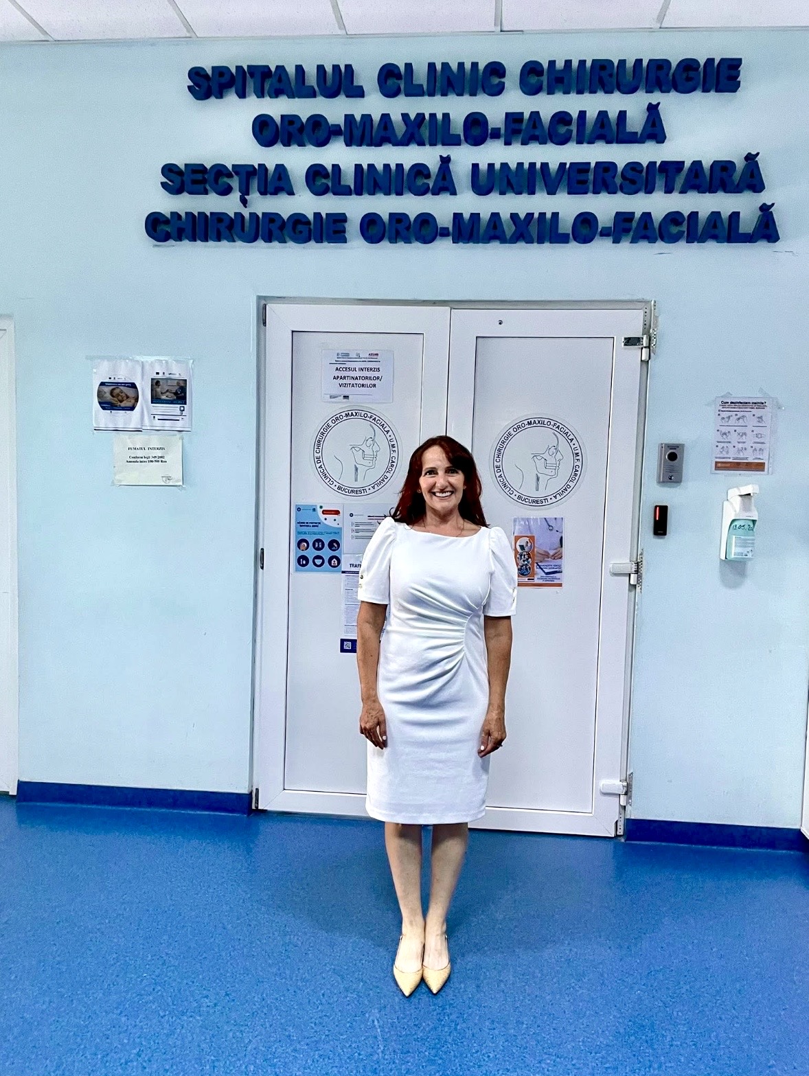 Woman in white dress standing in front of hospital doors with blue signage above, blue floor, and medical equipment nearby.