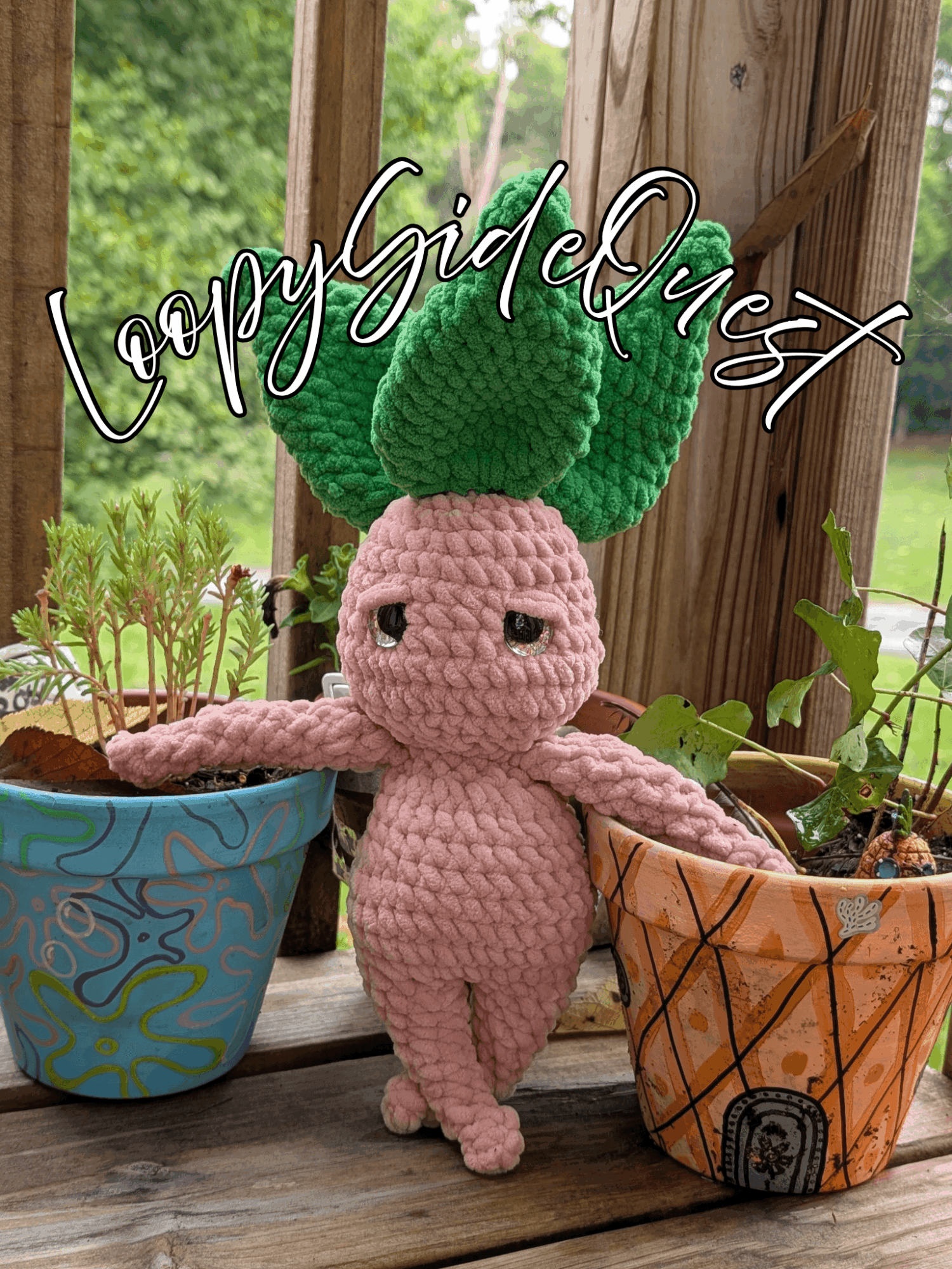 Pink crocheted creature with green leaves on head, standing between two colorful plant pots, with a wooden background and greenery.