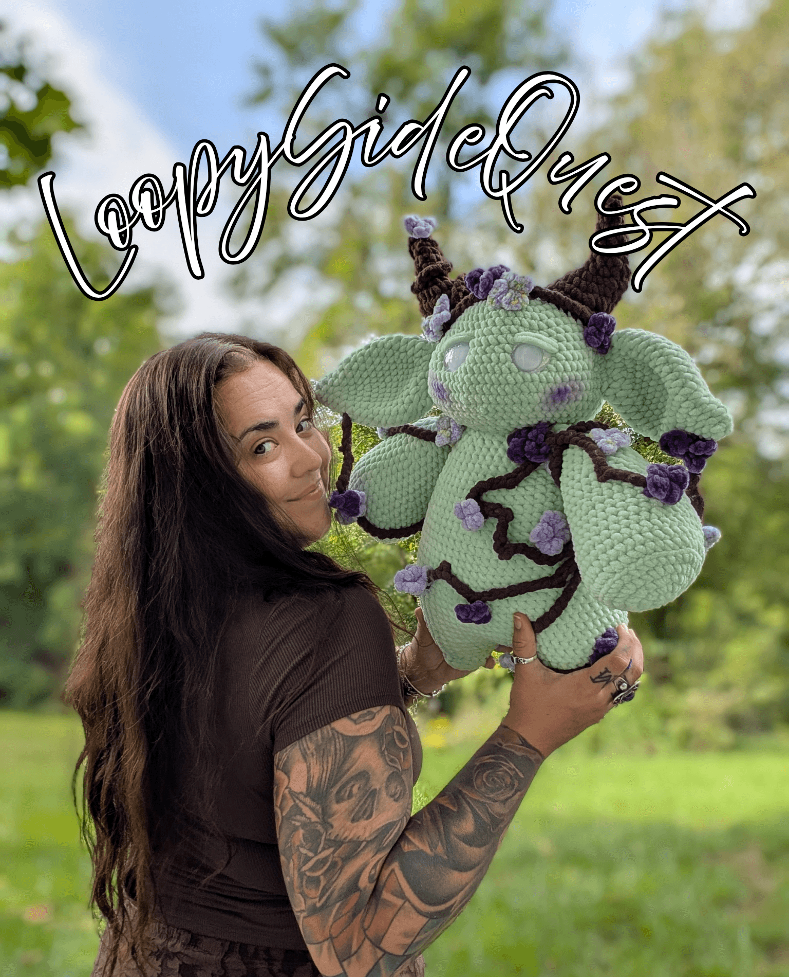 Woman with long dark hair and tattoos holding a large crocheted creature outdoors, with trees and blue sky in background.