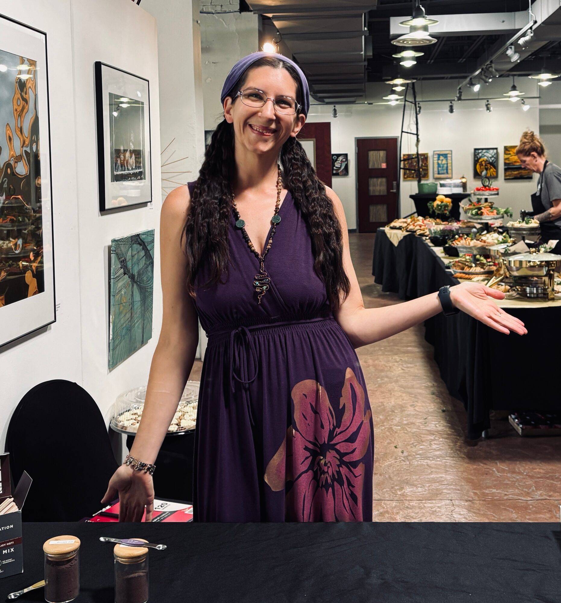 Smiling woman with long dark hair and glasses, wearing a purple dress with a large floral pattern, standing in an art gallery.