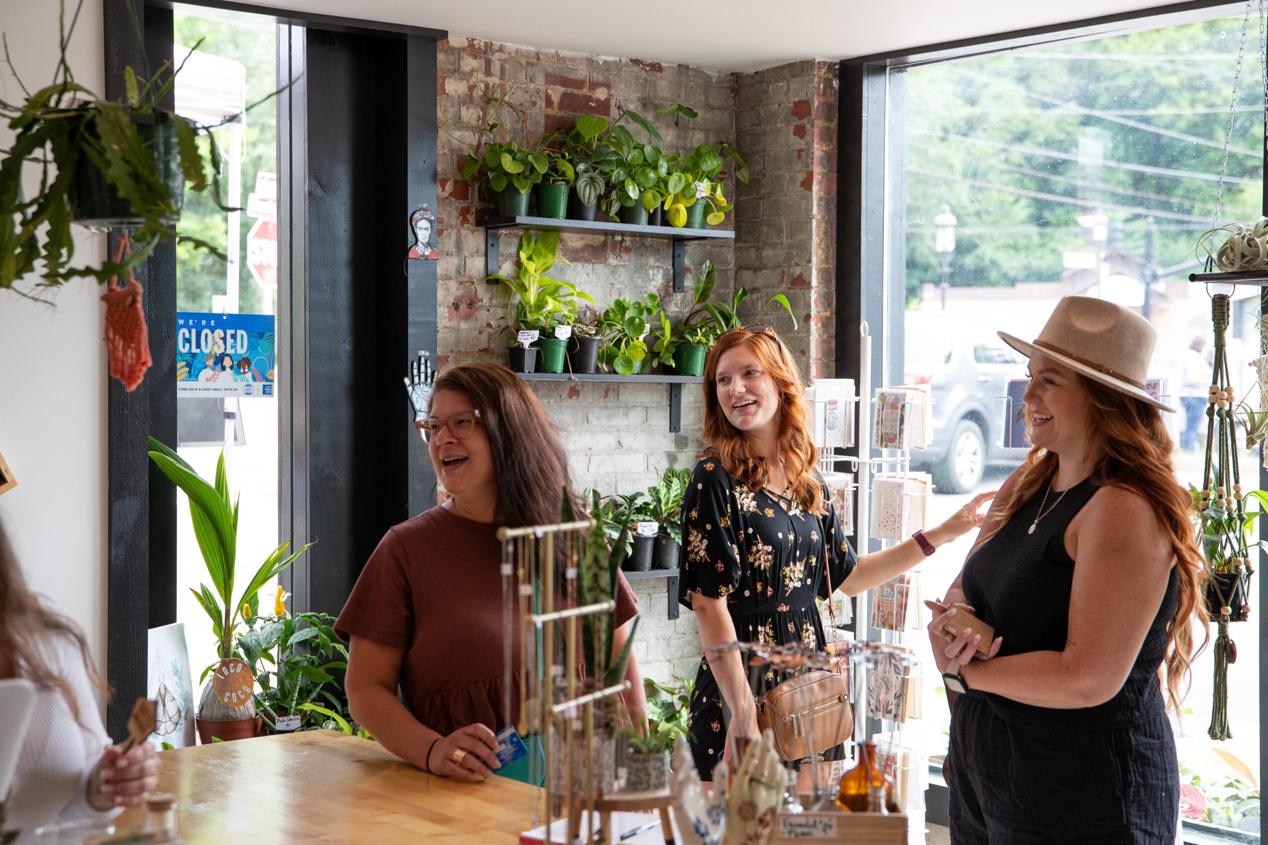 Three women are inside a plant shop, talking and smiling near a large window with plants on shelves and a brick wall.