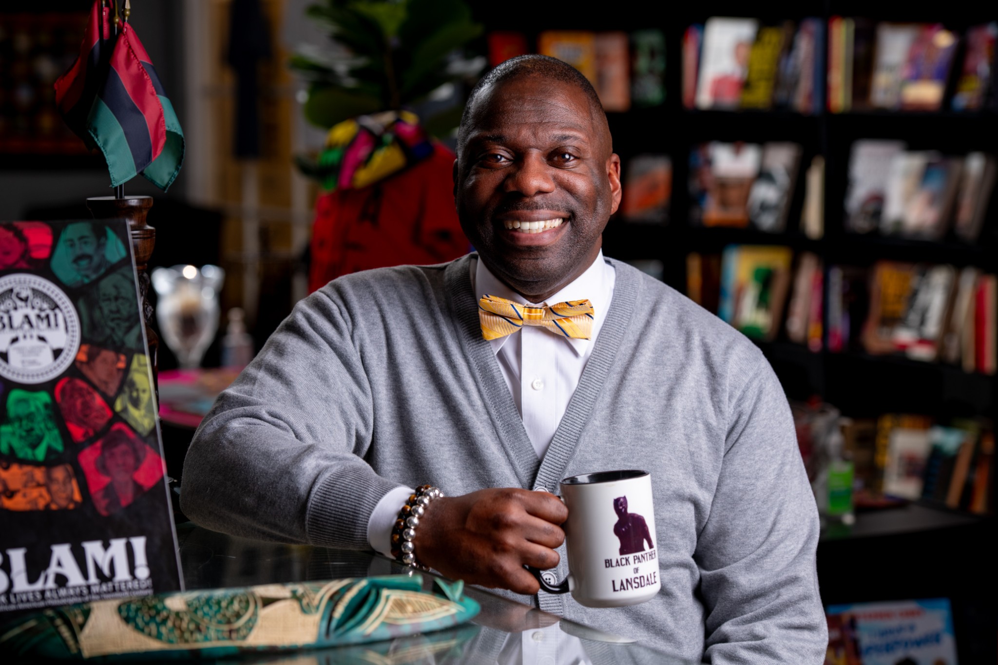 Smiling man with a yellow bow tie holding a mug in a bookstore or library setting.