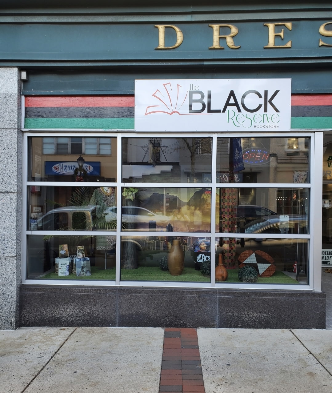 Store window display with various items, a sign reading 'BLACK RESCUE,' and reflections of street scene, sidewalk, and parked cars.