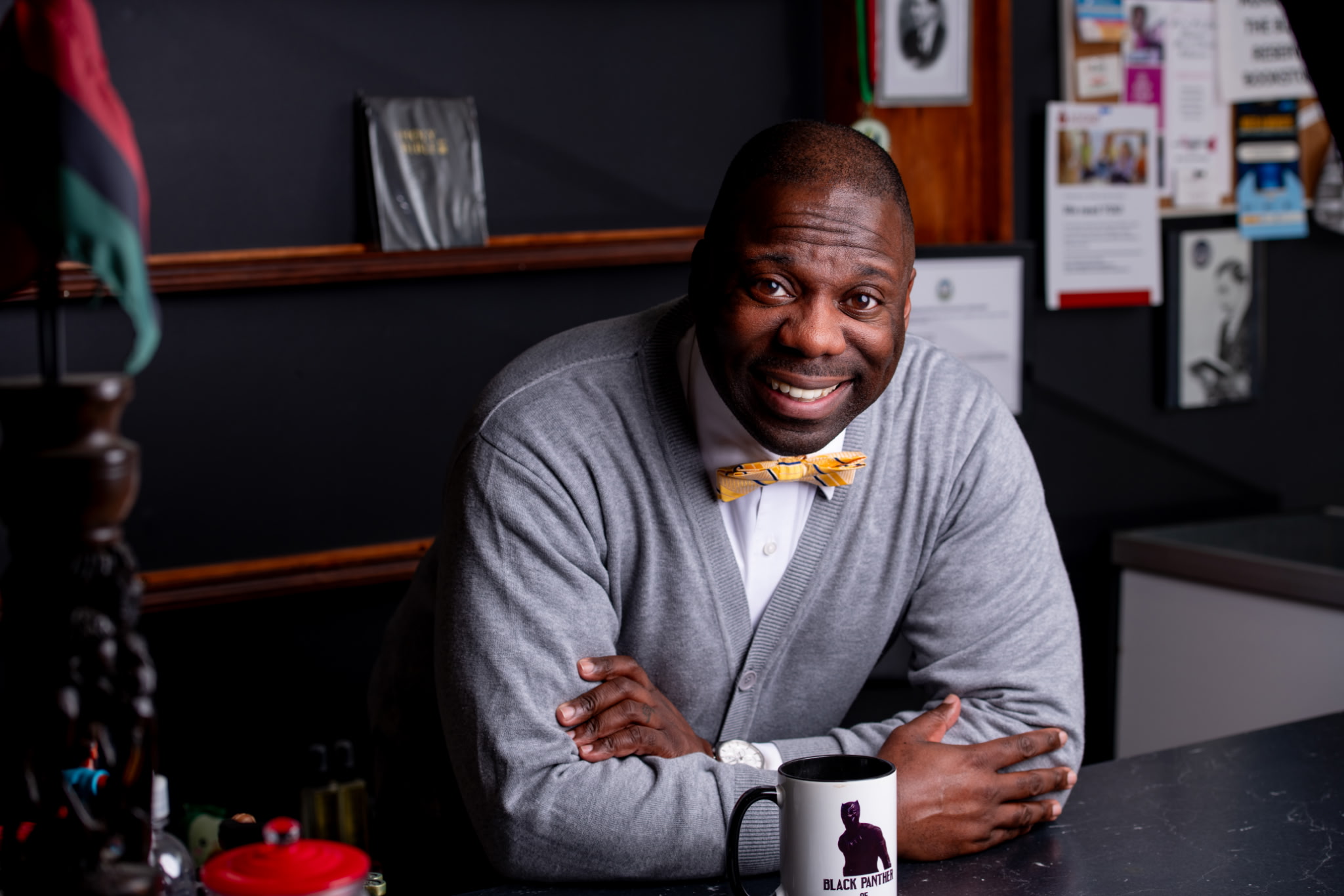 Man with a yellow bow tie smiling, sitting at a desk in an office, with bulletin boards behind him.