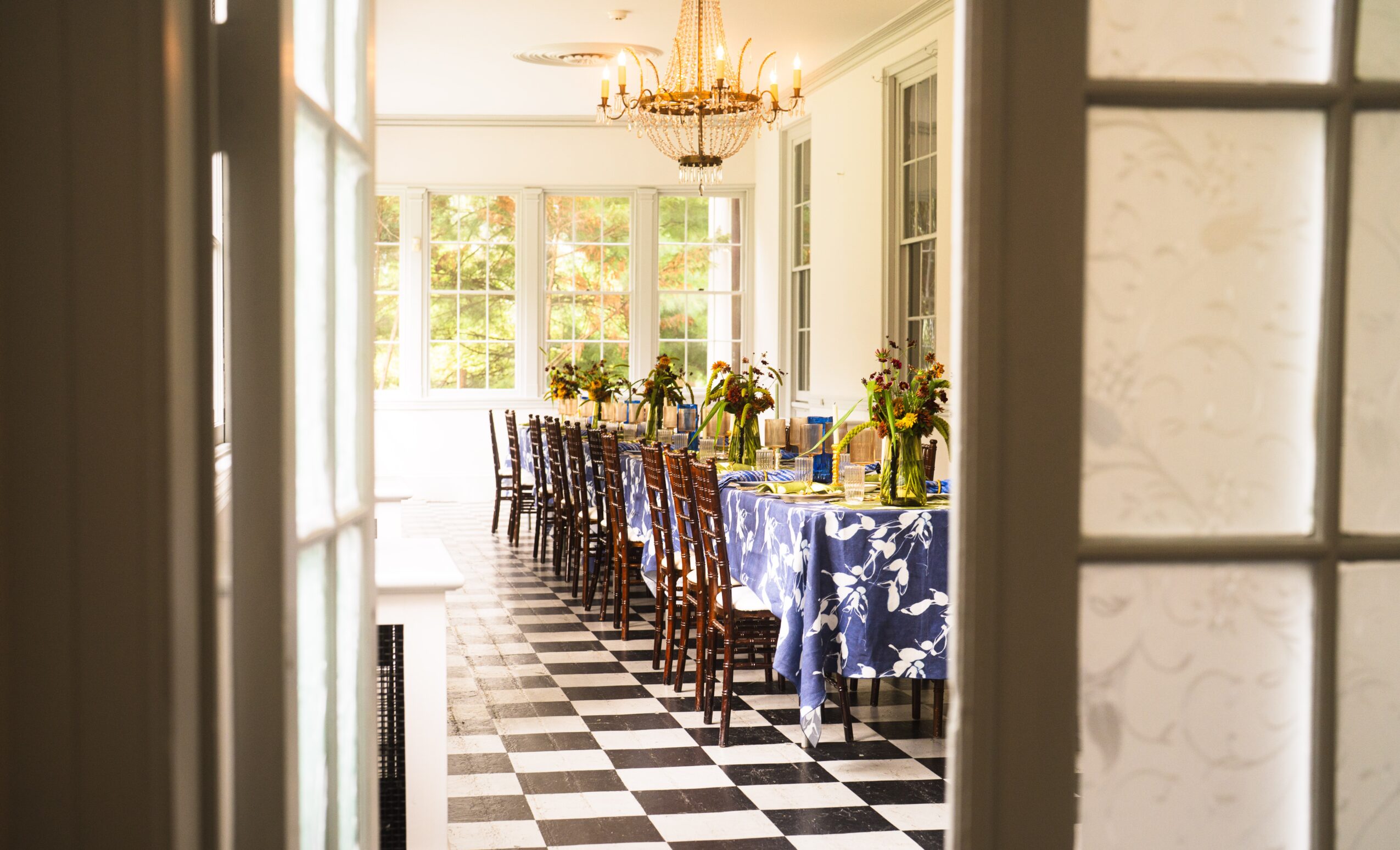 Dining room with long table, chairs, floral arrangements, checkered floor, and large windows, seen through a doorway.