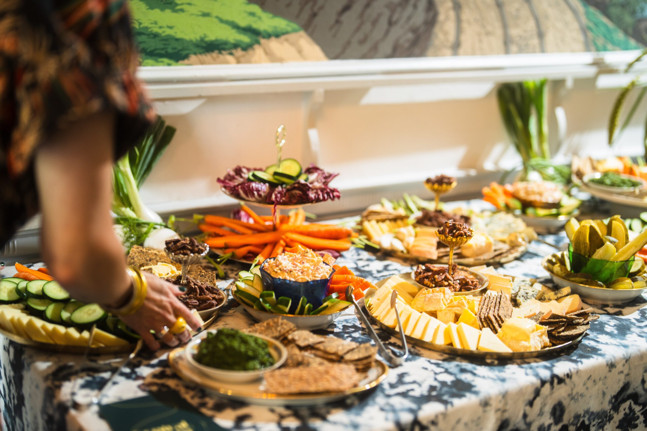 Table with various dishes and garnishes, person reaching for food, colorful presentation, outdoor setting, decorative tablecloth.
