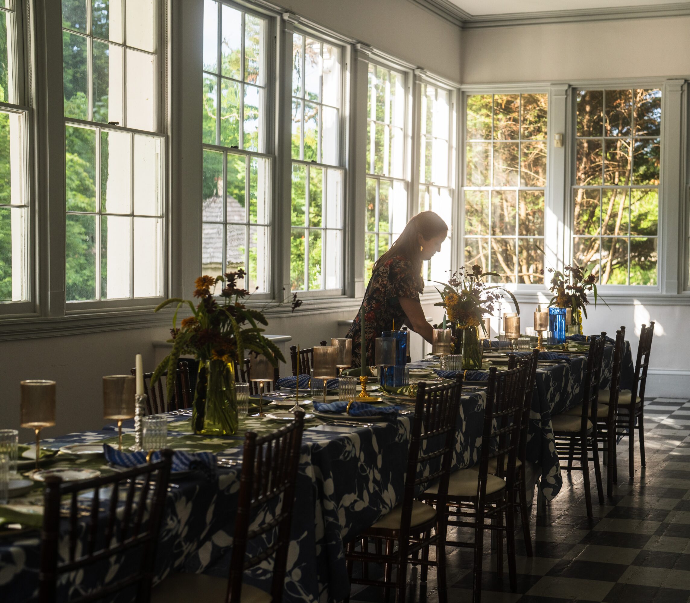 Person arranging flowers on a long dining table in a sunlit room with large windows and greenery outside.