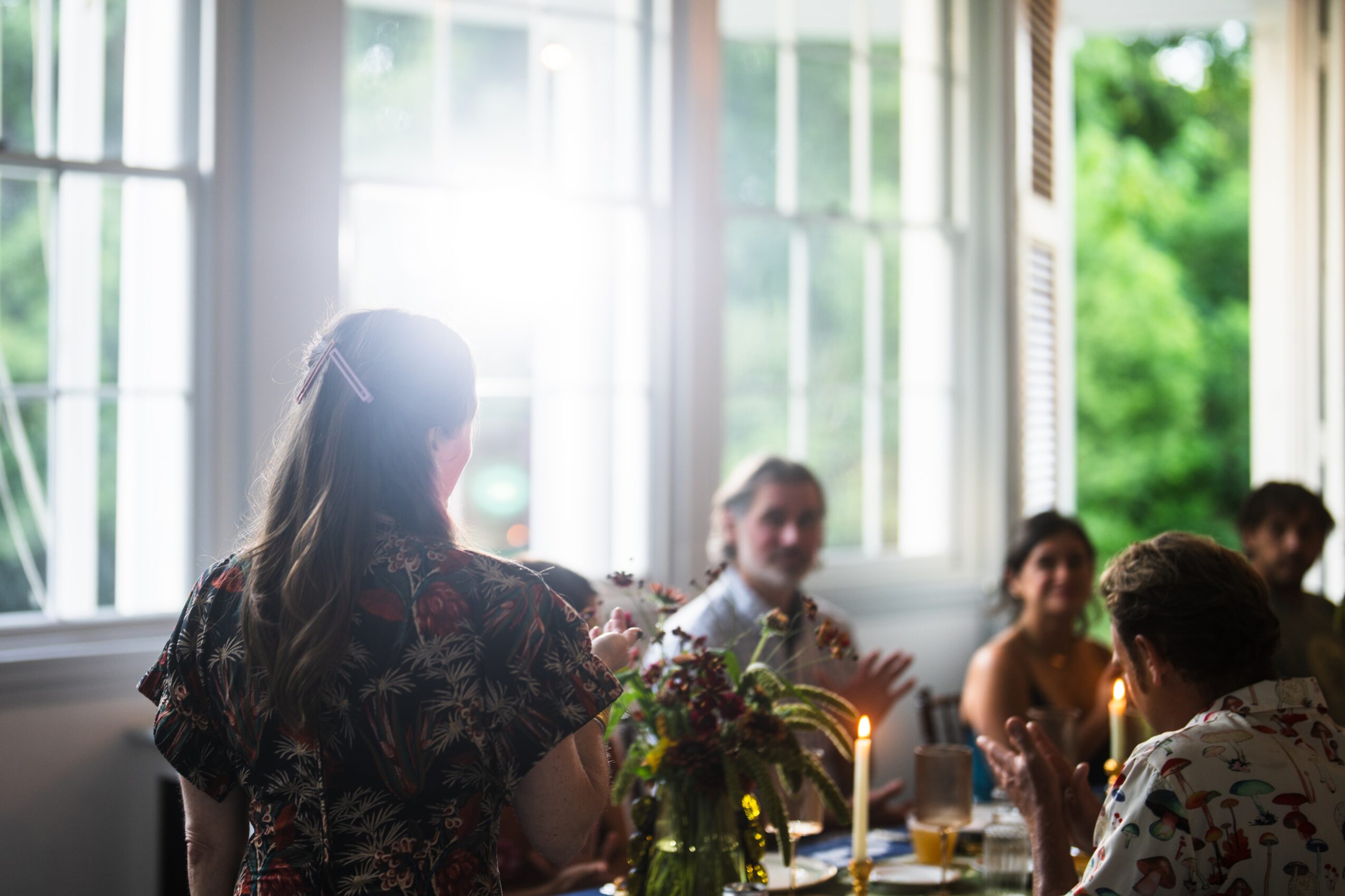 Group of people sitting at a table near large open windows with greenery outside, sunlight streaming in, woman standing with back to camera
