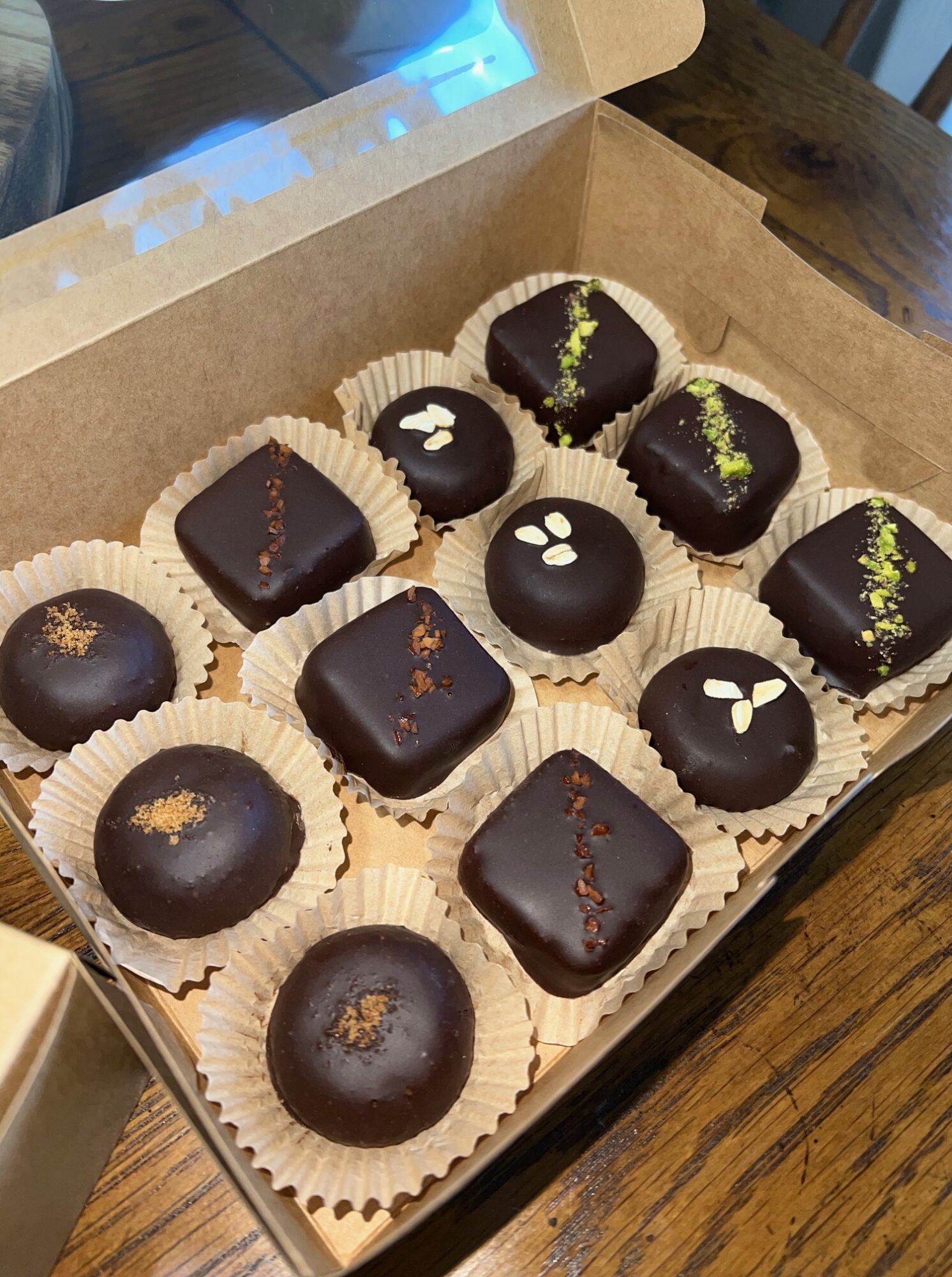 Box of assorted chocolates with various toppings and designs, arranged in a grid pattern on a wooden surface.