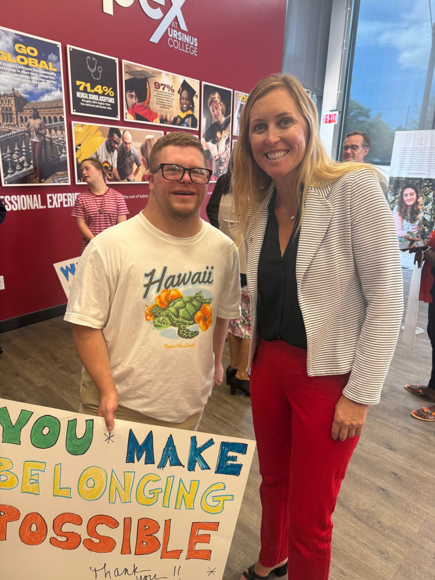 Two women standing indoors, one holding a colorful sign that reads 'You make lifelong impossible,' with posters and a window in the background.