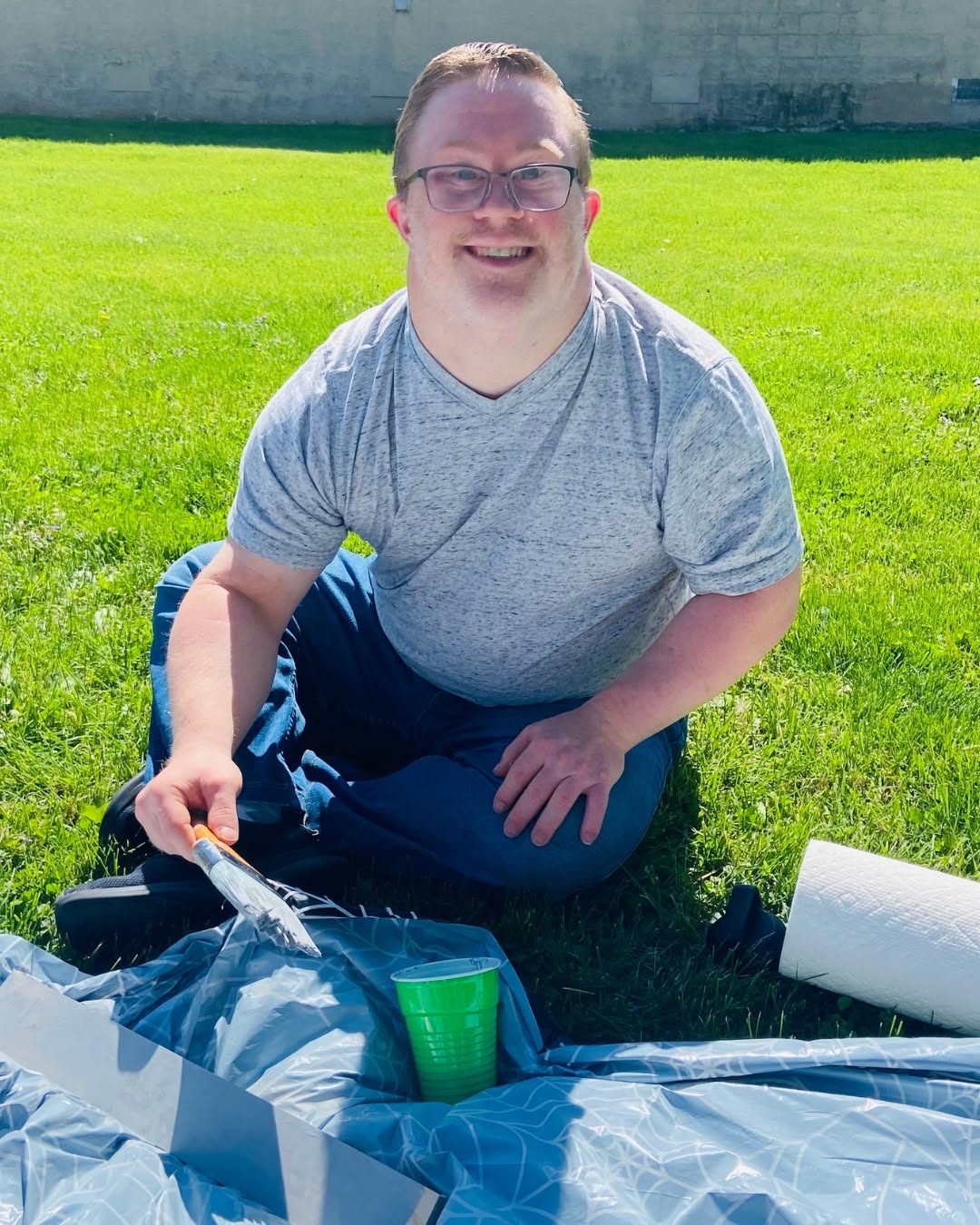 Person kneeling on grass outdoors, smiling, holding a tool, with a green cup nearby, bright sunny day.