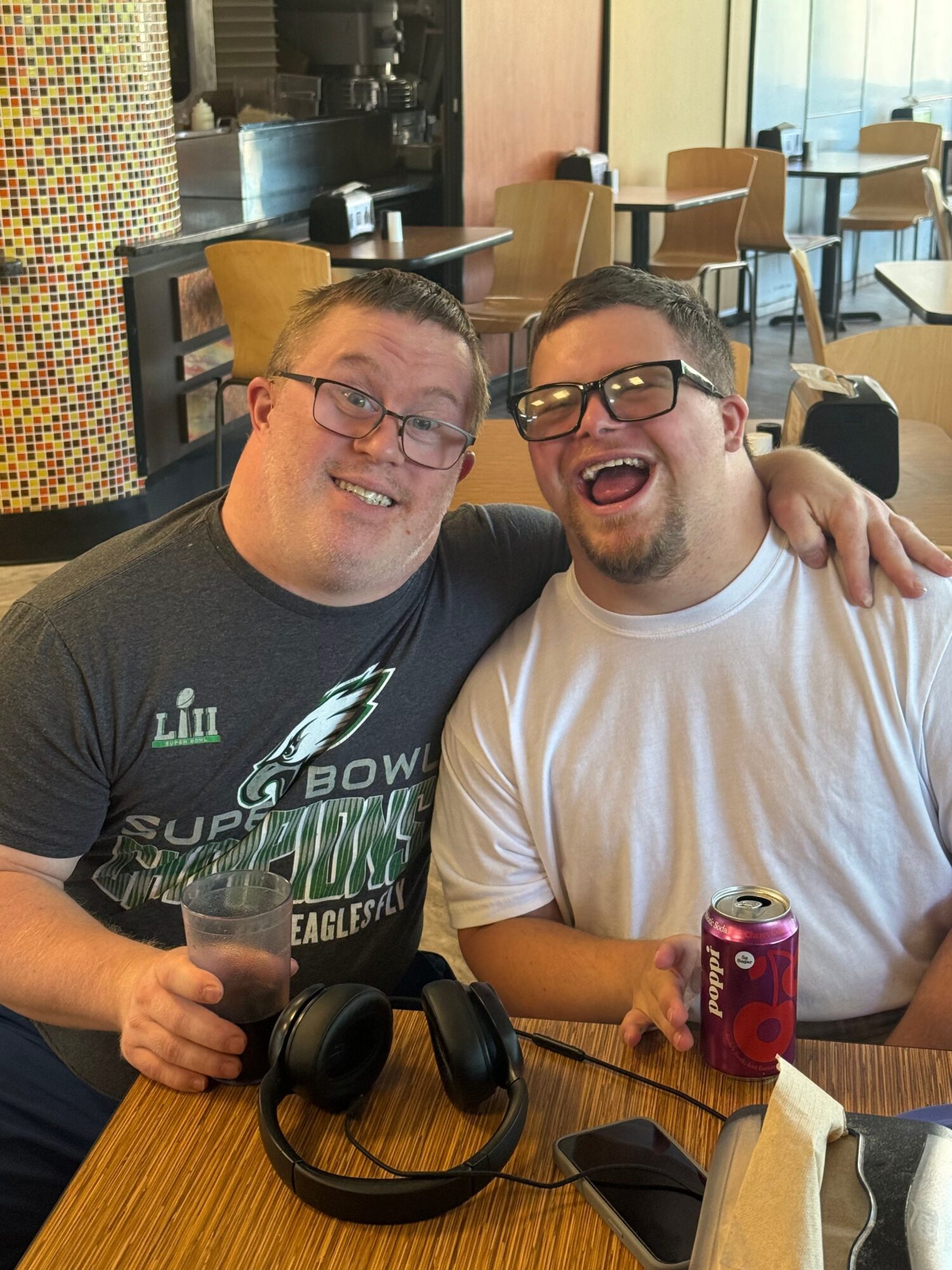 Two men with glasses sitting at a table, one holding a pink can, in a room with chairs and tables.