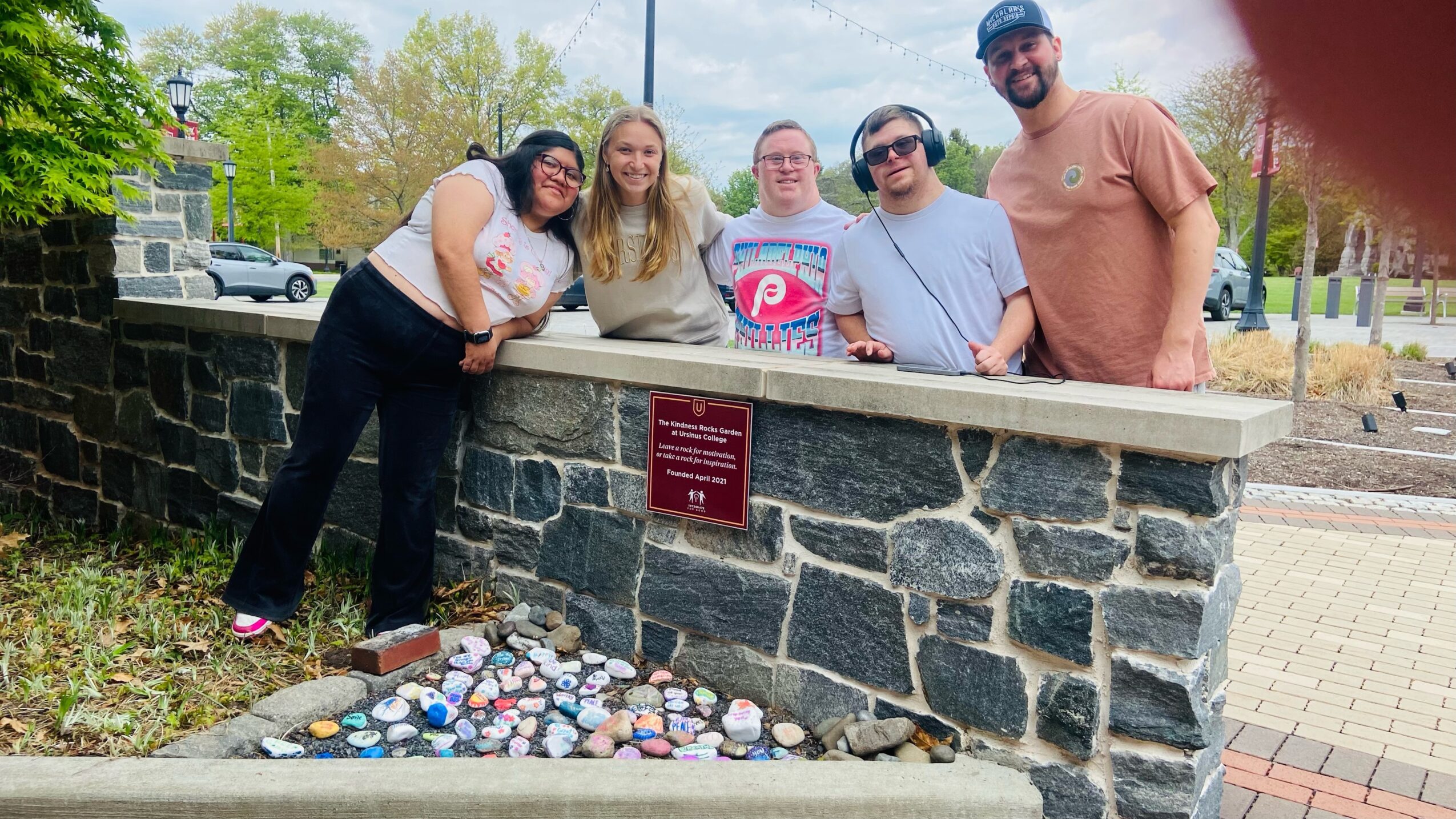 Group of five people leaning on a stone wall with a plaque, outdoors in a park-like setting.